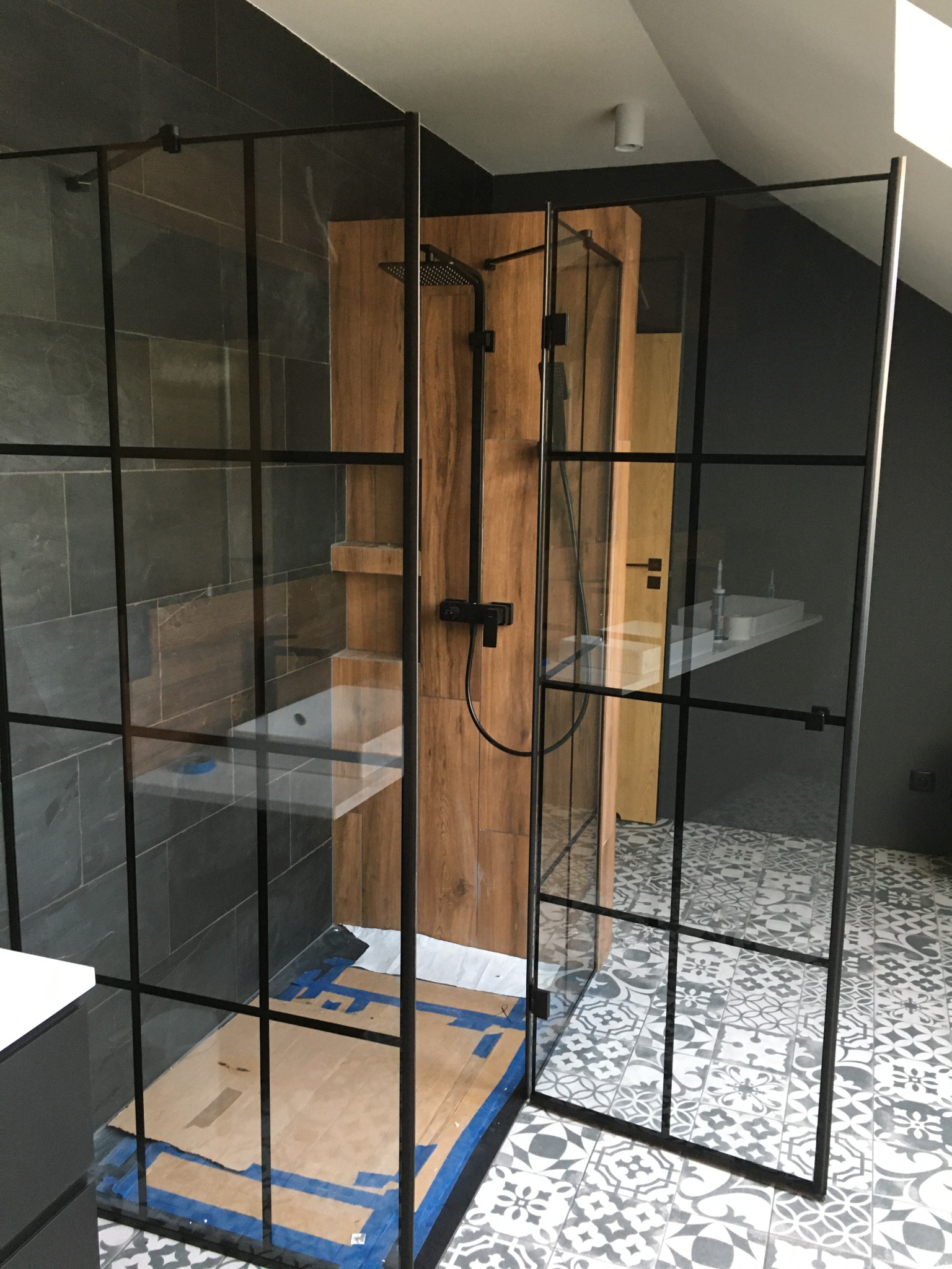 Nowoczesna kabina prysznicowa typu walk-in z czarnymi szprosami, drewnianym panelem prysznicowym i wzorzystą podłogą w łazience na poddaszu.