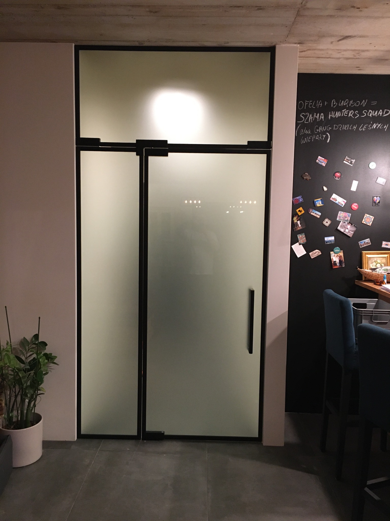 Szklane drzwi wewnętrzne z czarną ramą i matowym szkłem, przylegające do ściany z tablicą kredową i pamiątkami, obok doniczka z rośliną.