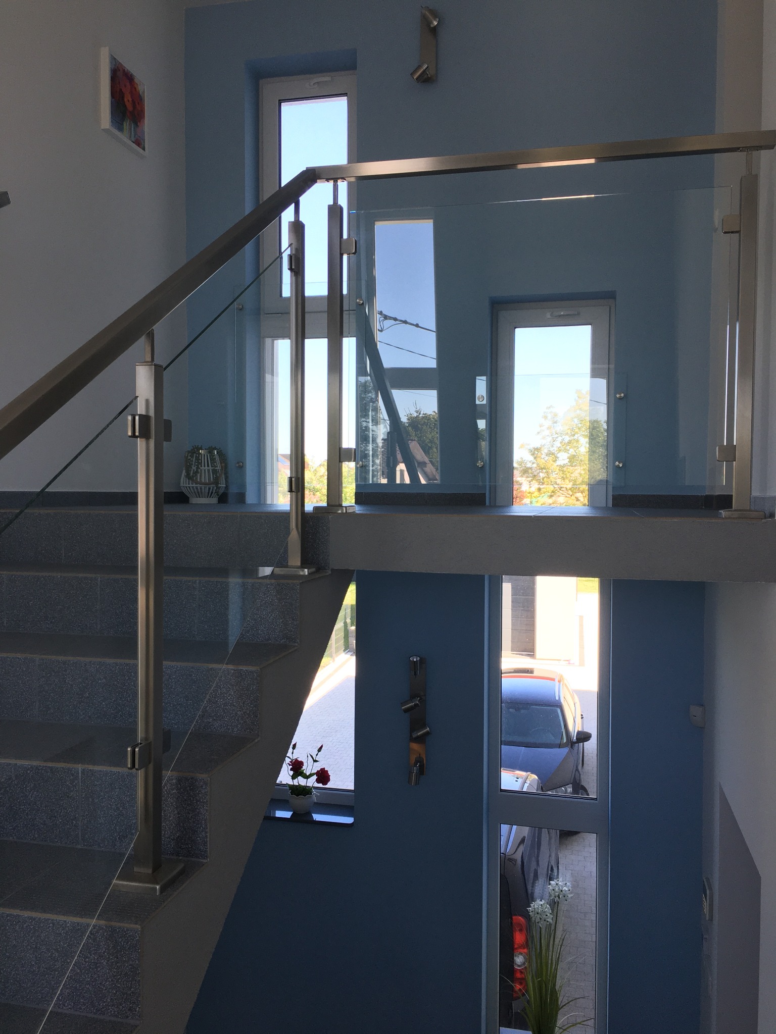 Wewnętrzna klatka schodowa z szarymi schodami i szklaną balustradą ze stali nierdzewnej, widok z góry na dół, kwiaty w doniczce na półpiętrze i przez okno widoczny samochód na zewnątrz.