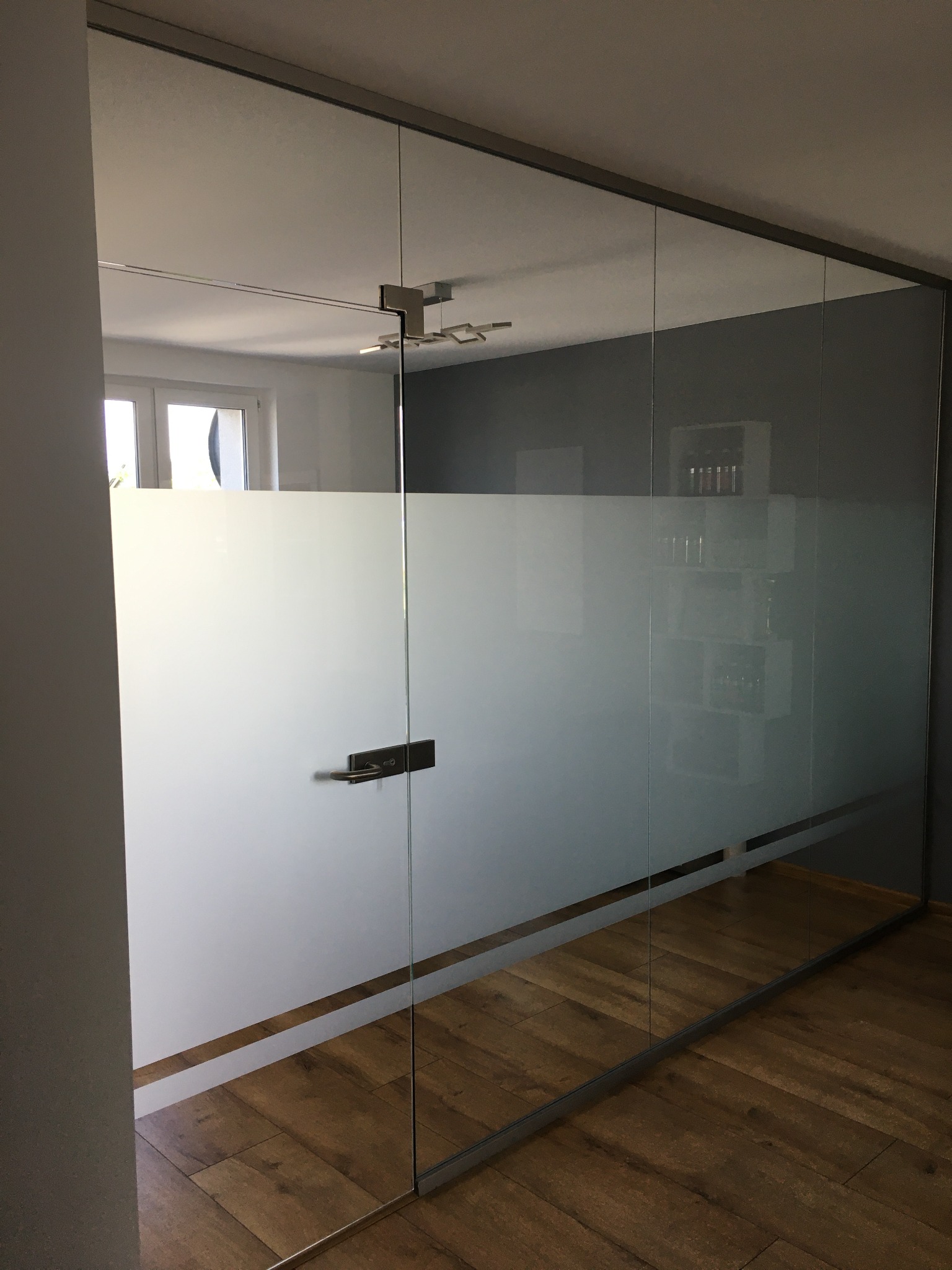 Szklana ściana działowa z częściowo matową folią, widoczna klamka i odblask drewnianej podłogi.