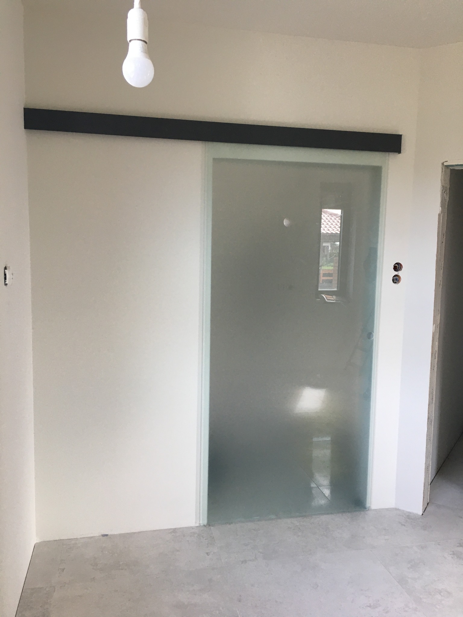 Zamontowane przesuwne drzwi szklane z czarną prowadnicą w surowym wnętrzu, widoczna żarówka bez klosza i fragment okna w odbiciu.