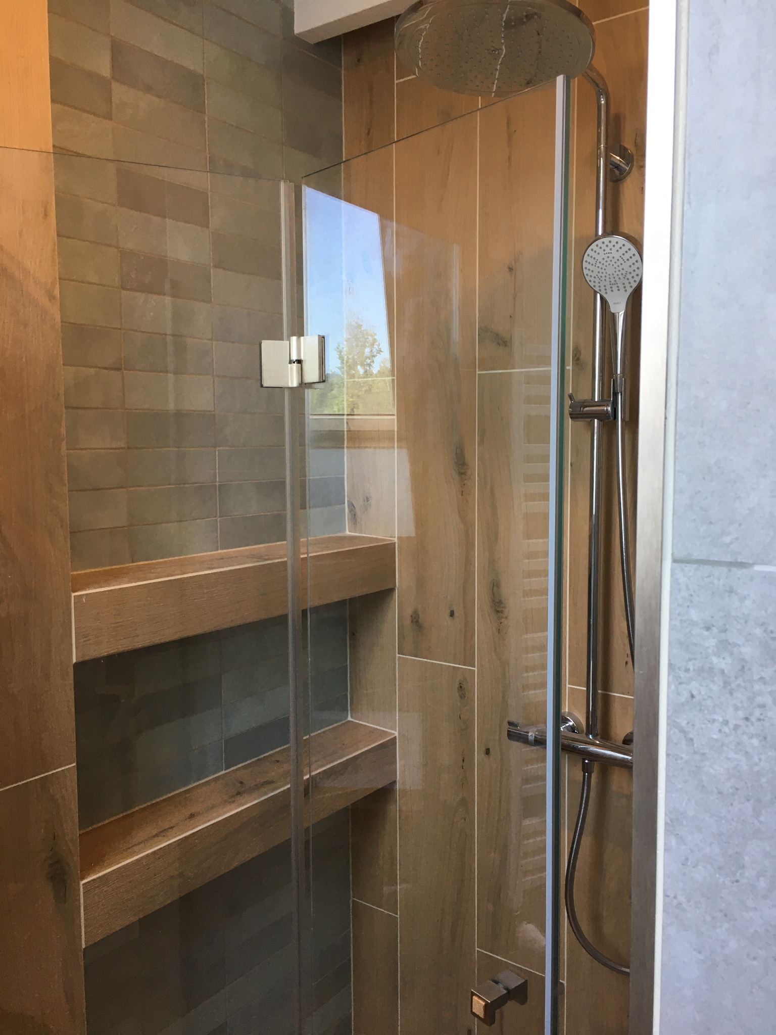 Kabina prysznicowa z szklanymi drzwiami, chromowaną armaturą i półkami wnękowymi wykończonymi drewnem, na tle płytek imitujących drewno.