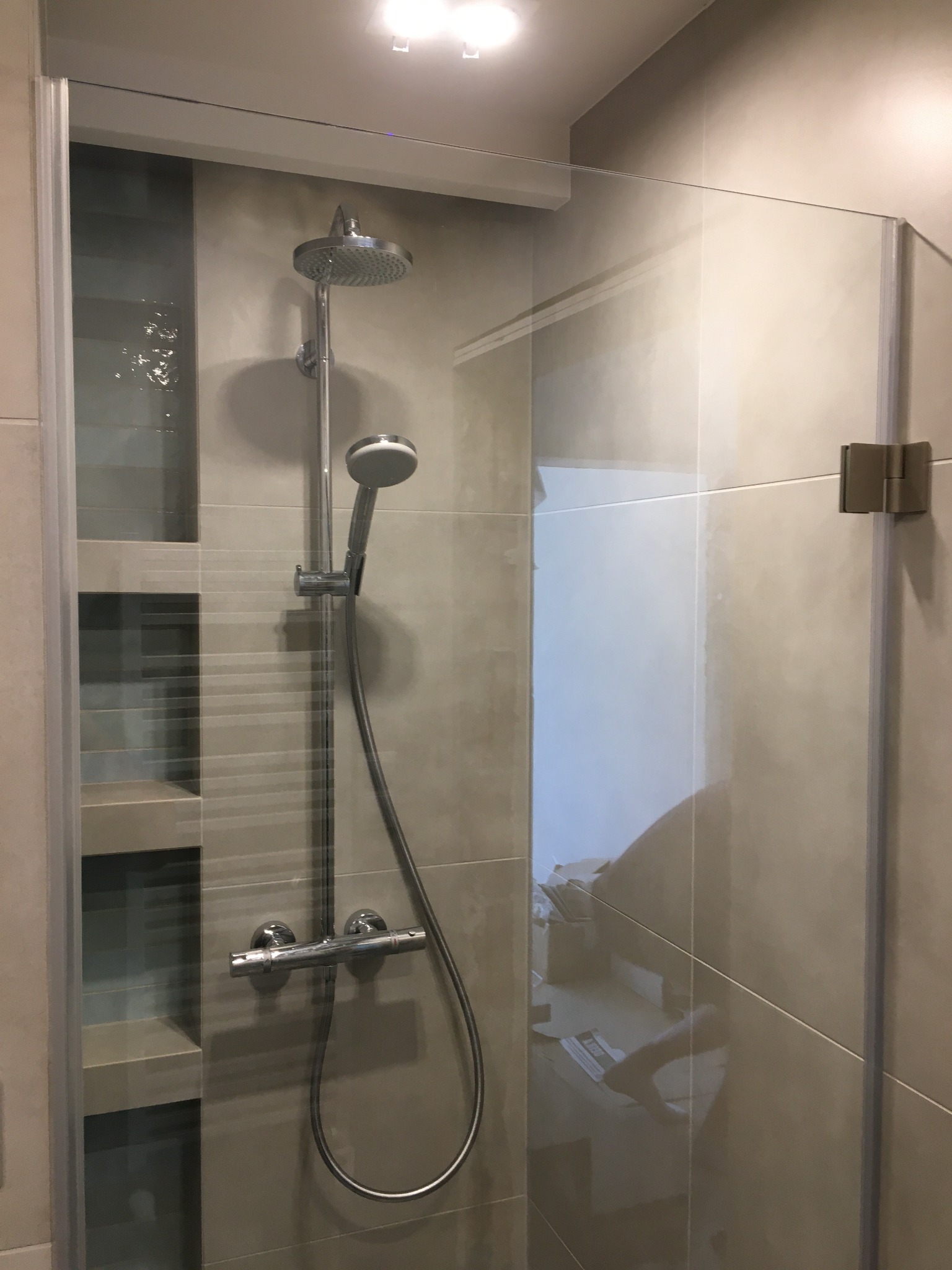 Szklana kabina prysznicowa z chromowaną armaturą i półkami wnękowymi, widoczne zawiasy mocujące szkło.