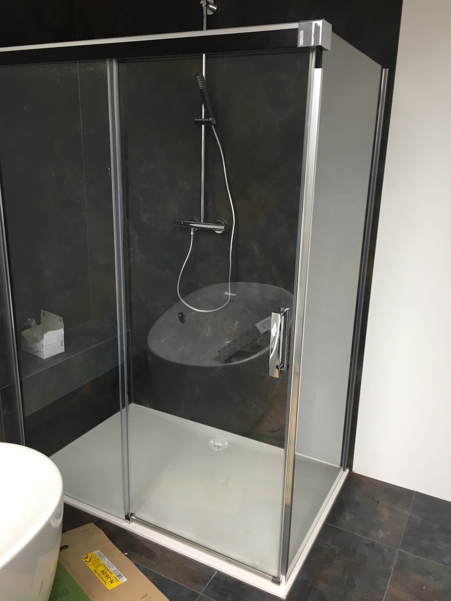 Kabina prysznicowa z przesuwnymi szklanymi drzwiami, zamontowana w łazience z ciemnymi płytkami i widoczną białą umywalką oraz pozostałościami po opakowaniu montażowym na podłodze.