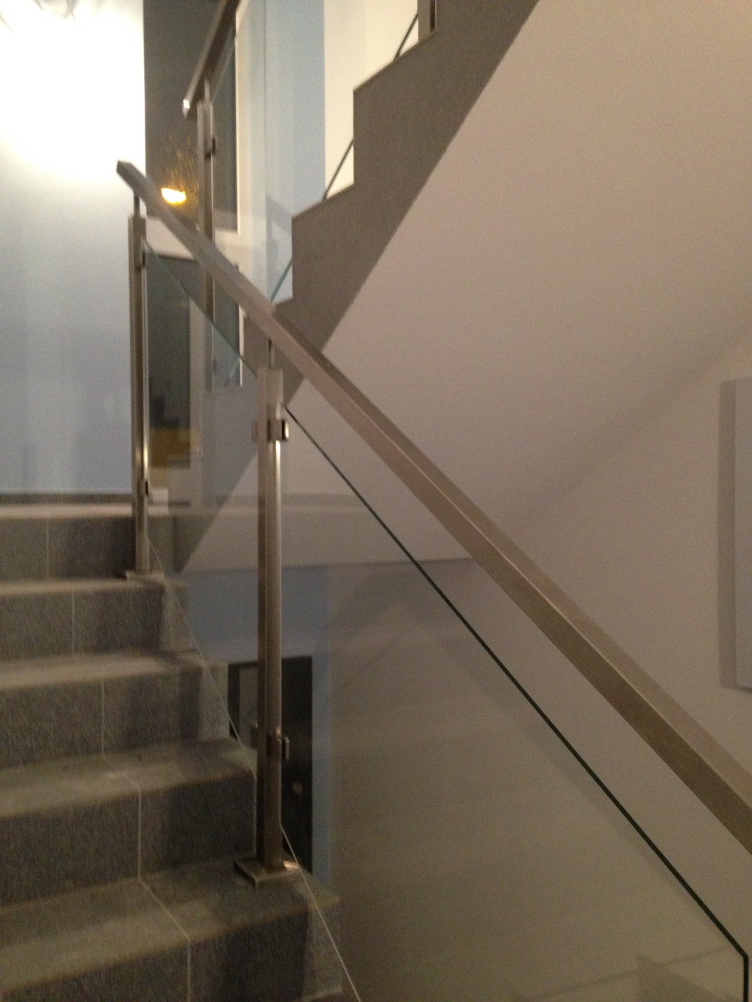 Szklana balustrada ze stalowymi słupkami zamontowana przy schodach wyłożonych szarą płytką, widok z dołu.