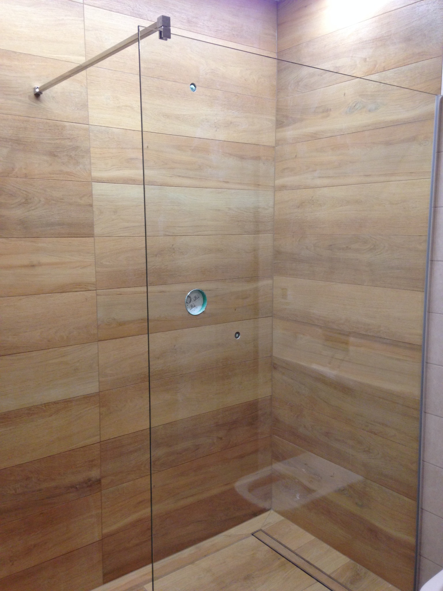 Kabina prysznicowa ze szklaną ścianką i drewnopodobnymi płytkami ceramicznymi na ścianach, widoczny metalowy uchwyt mocujący szybę.