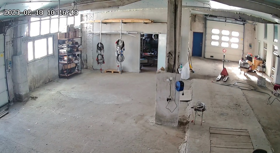 Przestronny magazyn z betonową podłogą, regałem w lewym rogu, zwisającymi kablami i wózkiem paletowym w tle, oświetlony naturalnym światłem wpadającym przez okna.