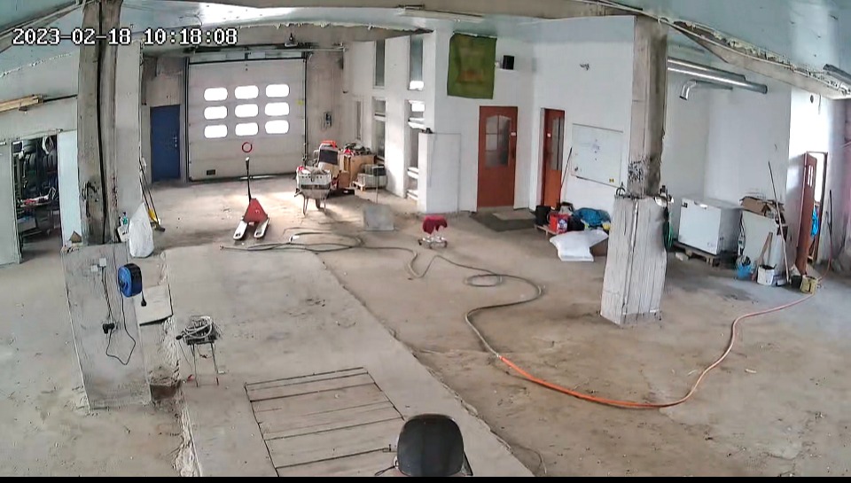 Wnętrze magazynu lub hali z bramą garażową, wózkiem paletowym, narzędziami i przewodami na podłodze.