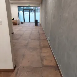 Wnętrze biura w trakcie wykańczania, widoczne brązowe płytki podłogowe, surowa szara ściana z widocznymi otworami, kartony i sprzęt w tle, oraz przeszklone drzwi wejściowe.