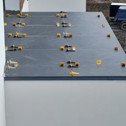 Układanie ciemnych płytek na balkonie z użyciem żółtych klinów poziomujących, widoczny fragment ściany budynku i ciężarówka w tle.