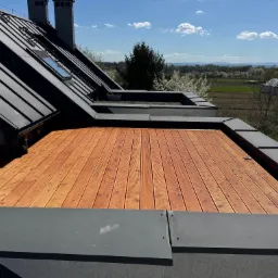 Nowoczesny taras drewniany na dachu w Nowym Rybiu. Widok na okolicę, dach pokryty blachą. Deski tarasowe ułożone równolegle, szara obudowa.