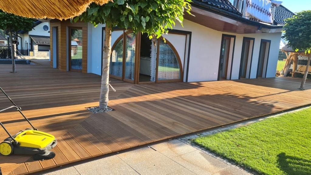 Rozległy taras z desek kompozytowych, z wbudowanymi drzewami i widokiem na dom z zaokrąglonymi oknami. Żółta zamiatarka do liści na pierwszym planie.