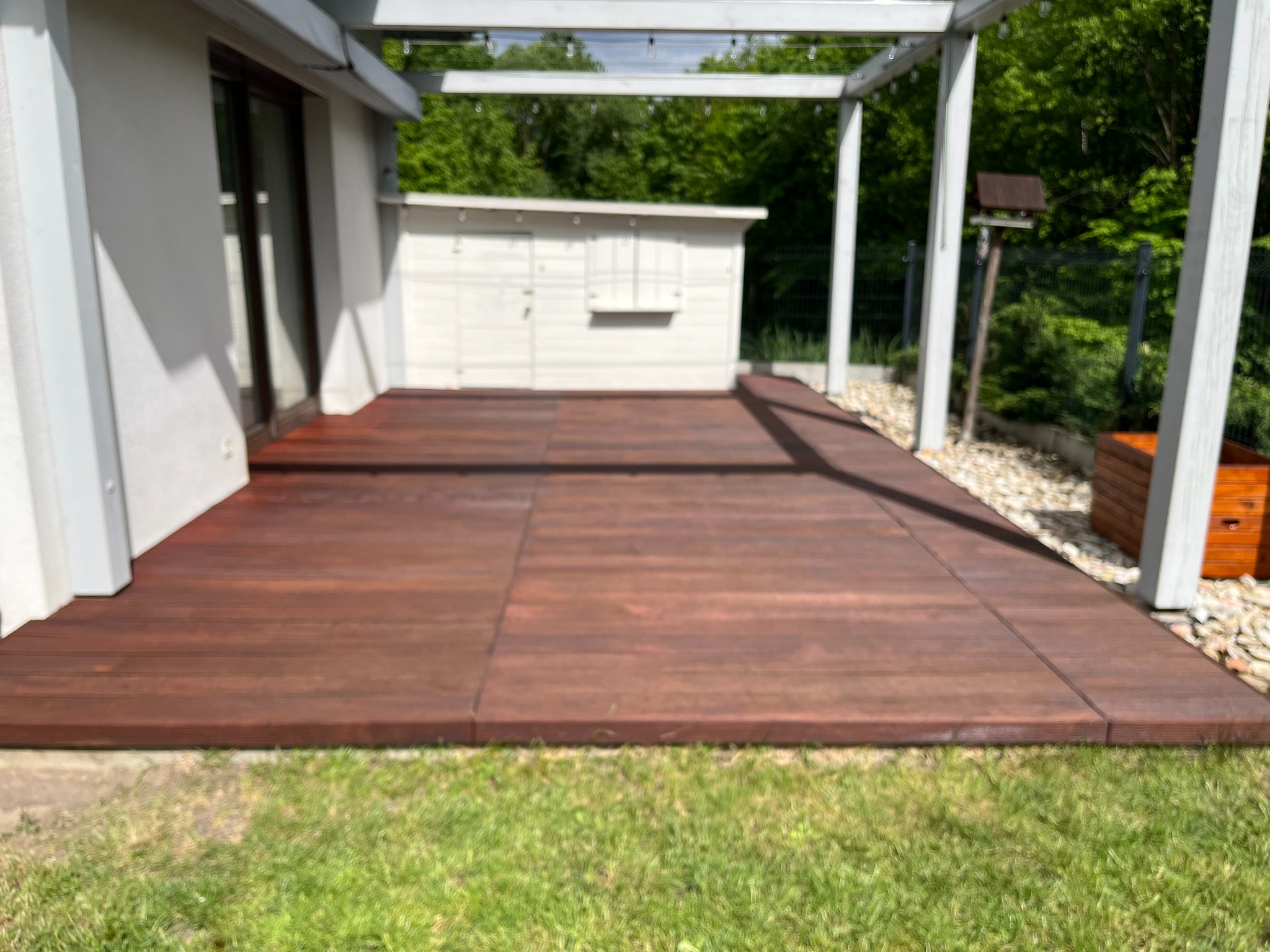 Ciemnobrązowy taras z drewna egzotycznego, zadaszony pergolą, przylegający do białego budynku. W tle zieleń drzew i krzewów, widoczny fragment trawnika.