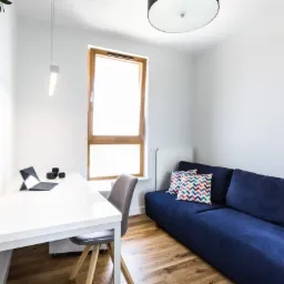 Pokój z białym biurkiem, laptopem, ciemnoniebieską sofą i drewnianą podłogą, oświetlony wiszącą lampą i światłem z okna.