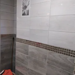 Wykończona ściana łazienki z szarymi i białymi płytkami imitującymi drewno, dekorowana mozaiką z brązowych kostek, z czarno-białą grafiką kwiatu na górze i elementami odzieży w rogu.