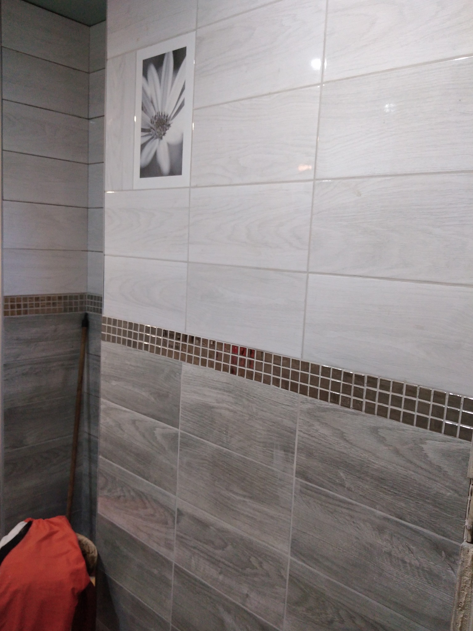 Wykończona ściana łazienki z szarymi i białymi płytkami imitującymi drewno, dekorowana mozaiką z brązowych kostek, z czarno-białą grafiką kwiatu na górze i elementami odzieży w rogu.