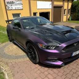 Fioletowy Ford Mustang z czarną maską stoi przed warsztatem Mar-Gaz, specjalizującym się w montażu i serwisie instalacji LPG. Widoczne panele słoneczne na dachu budynku.