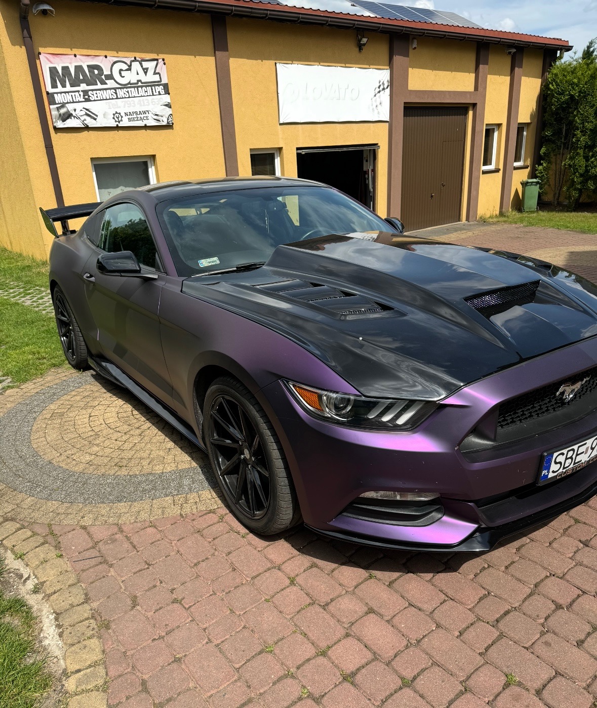 Fioletowy Ford Mustang z czarną maską stoi przed warsztatem Mar-Gaz, specjalizującym się w montażu i serwisie instalacji LPG. Widoczne panele słoneczne na dachu budynku.