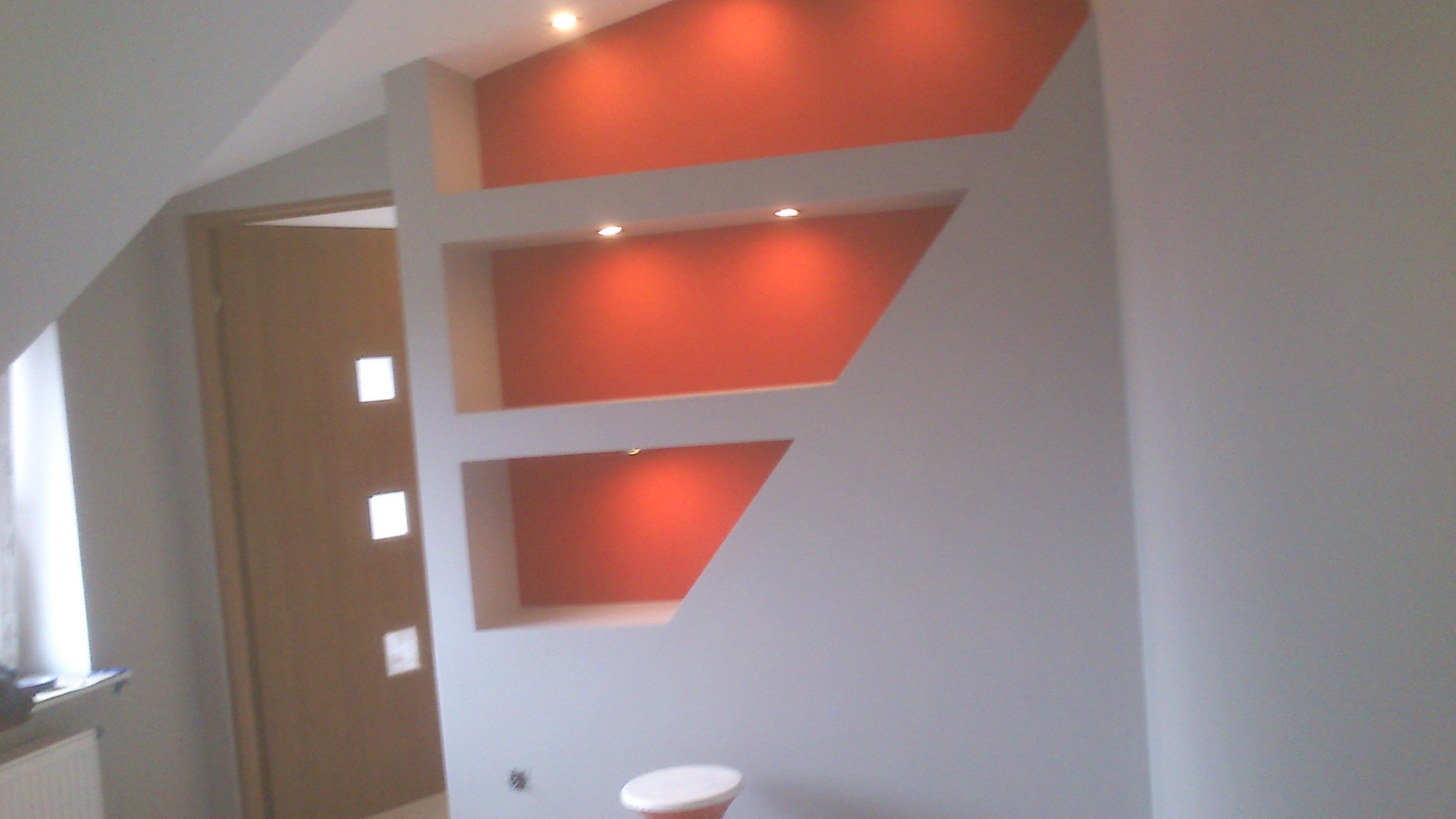 Ściana w pokoju z nieregularnymi wnękami pomalowanymi na pomarańczowo, oświetlonymi punktowo, widoczny fragment drzwi z kwadratowymi okienkami i fragment stołka.