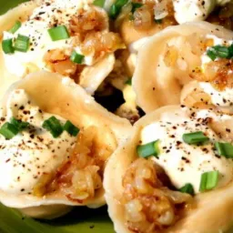 Porcja pierogów z serem, polanych śmietaną, skwarkami z cebuli i posypanych zielonym szczypiorkiem, podana na zielonym talerzu.