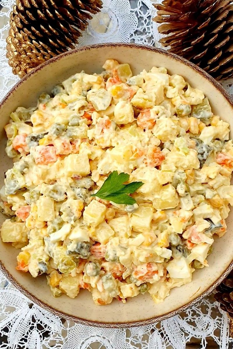 Świąteczna sałatka jarzynowa w beżowej misce, udekorowana gałązką pietruszki, na tle białej koronki i złotych szyszek. Idealna propozycja na catering.