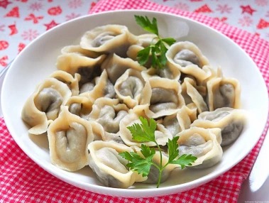 Porcja pierogów na białym talerzu, dekorowana świeżą natką pietruszki. Tło stanowi czerwono-biała krata, sugerująca domowy klimat. Delikatne oświetlenie podkreśla świeżość dania.