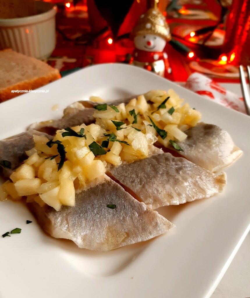 Śledź w oleju z jabłkiem i cebulką, posypany zieloną pietruszką, na białym talerzu. W tle dekoracje świąteczne i pieczywo. Tradycyjna przekąska na stole.