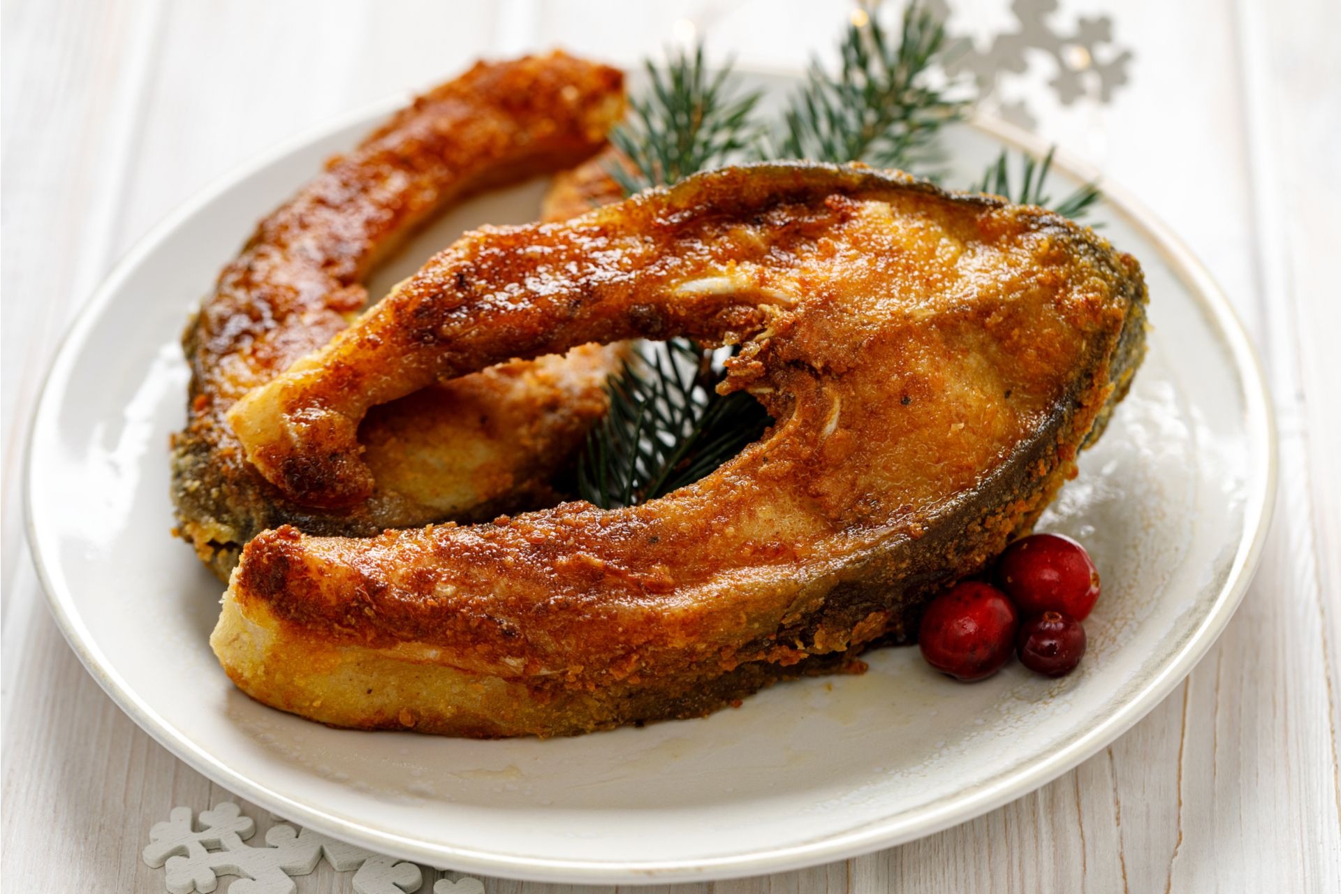 Dwa smażone kawałki karpia na białym talerzu, udekorowane gałązką świerku i żurawiną, na tle białych desek i ozdoby w kształcie śnieżynki.
