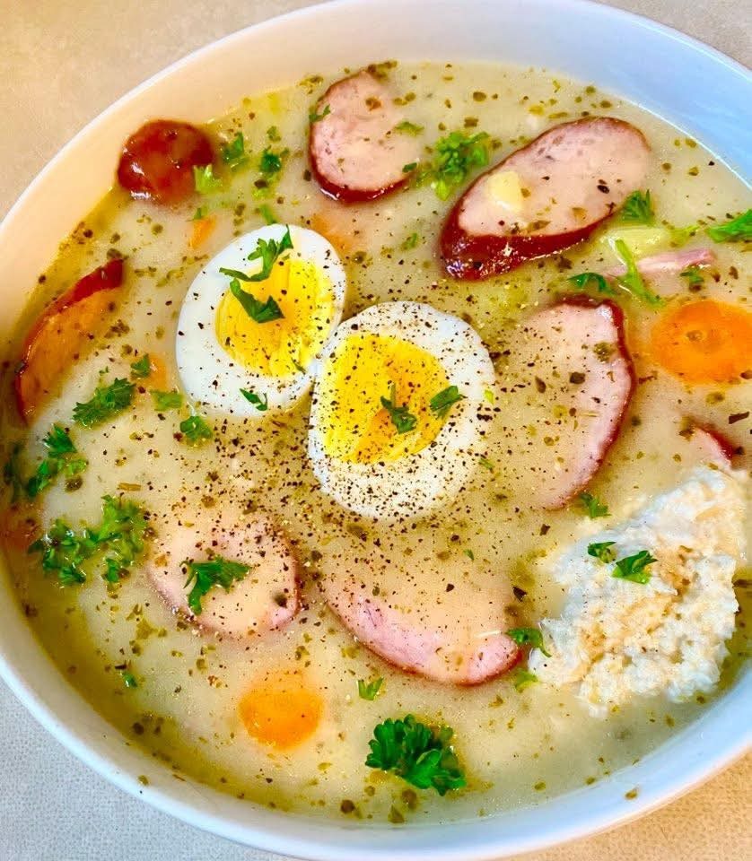 Żurek w białej misce, udekorowany plasterkami jajka, kiełbasy i świeżą natką pietruszki. Zupa doprawiona pieprzem, widoczne kawałki warzyw. Tradycyjne danie kuchni polskiej.