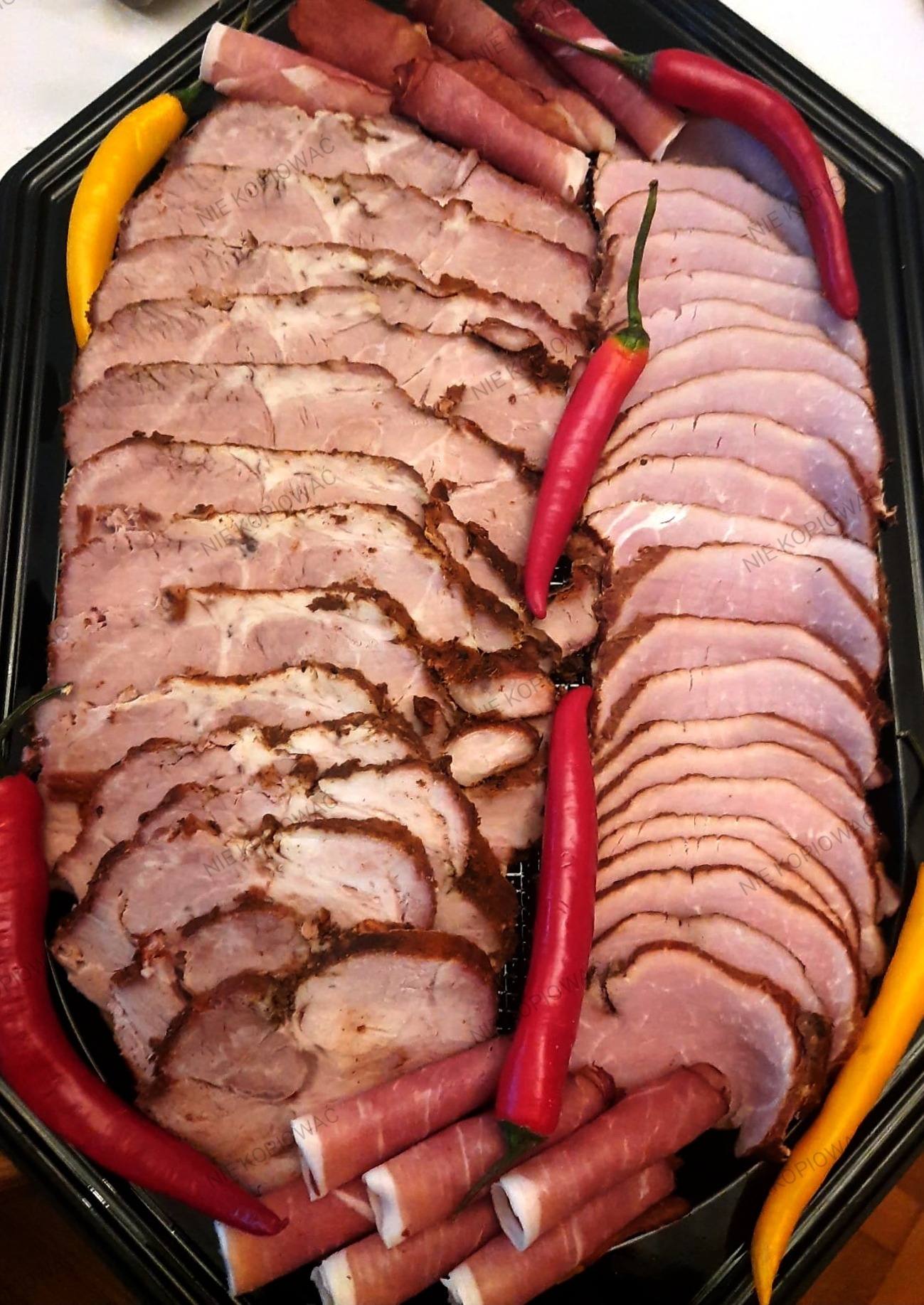 Elegancka taca z plastrami pieczonego mięsa, szynki zwiniętej w rulony i papryczek chili w kolorach czerwonym i żółtym, tworząca apetyczną kompozycję.