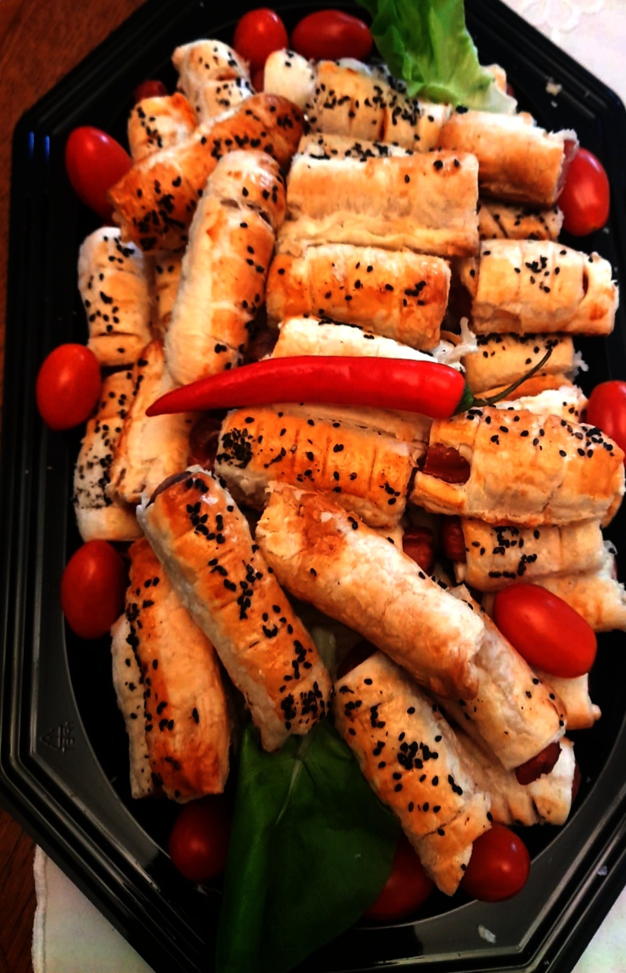 Apetyczne koreczki z ciasta francuskiego z parówką, posypane czarnuszką, ułożone na czarnym talerzu z pomidorkami koktajlowymi i papryczką chili. Idealne na catering.