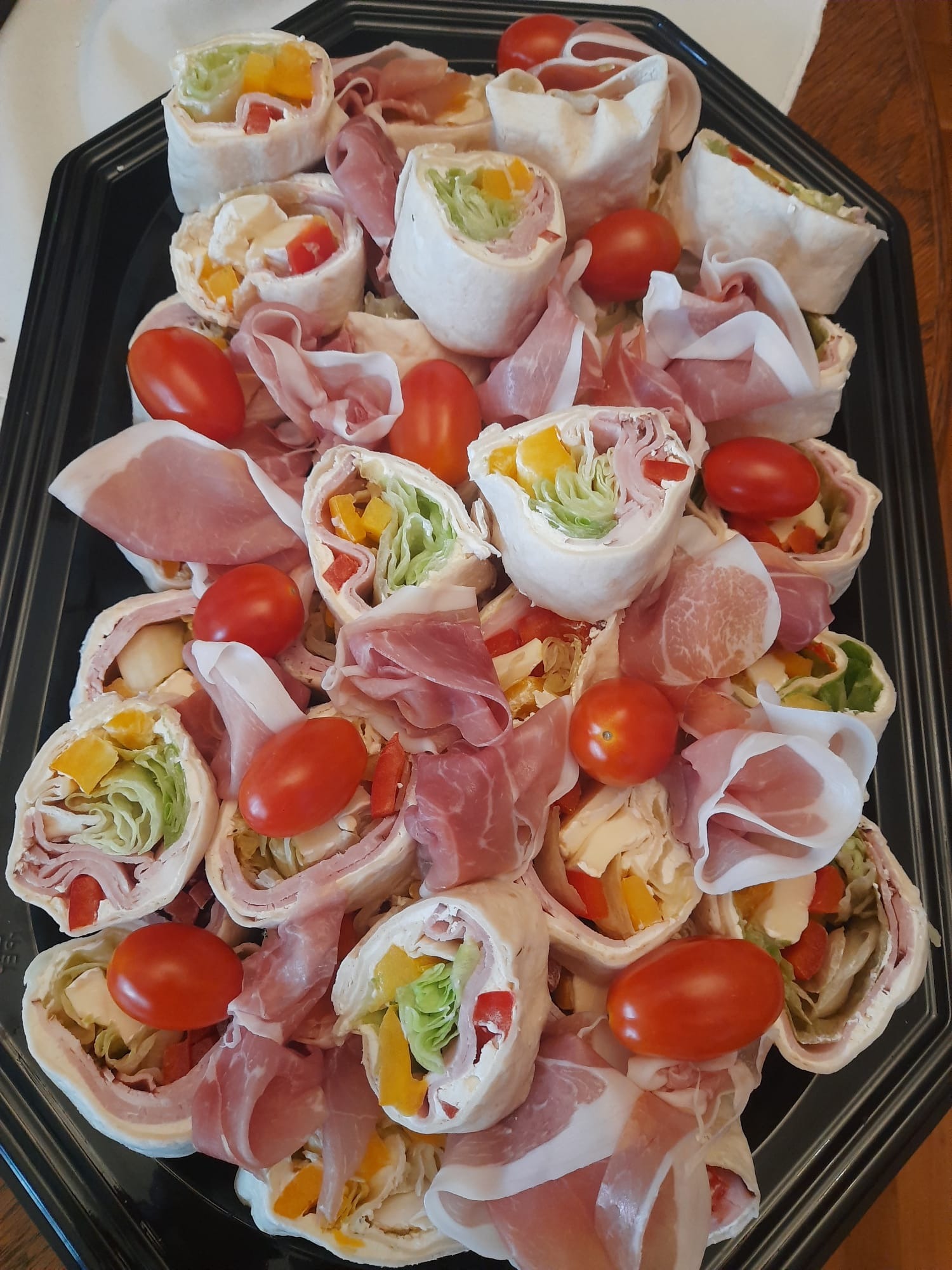 Tortilla wrap rolls z szynką parmeńską i pomidorkami koktajlowymi na czarnej tacy. Widoczne kolorowe warzywa: sałata, papryka. Idealne na catering firmowy.