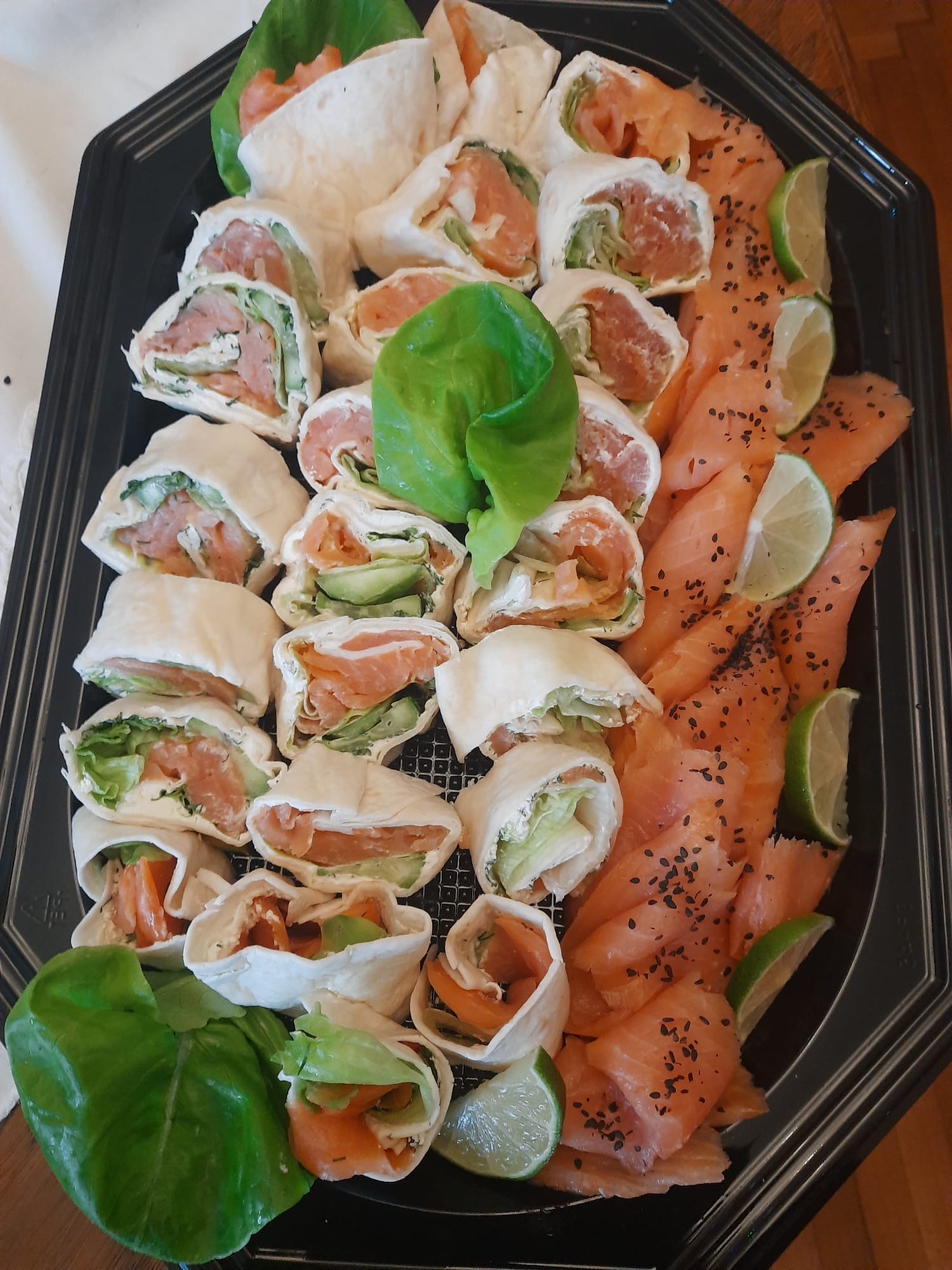 Elegancka taca z roladkami z łososiem, plastrami łososia i limonką, dekorowana zielonymi liśćmi sałaty. Idealna propozycja na catering.