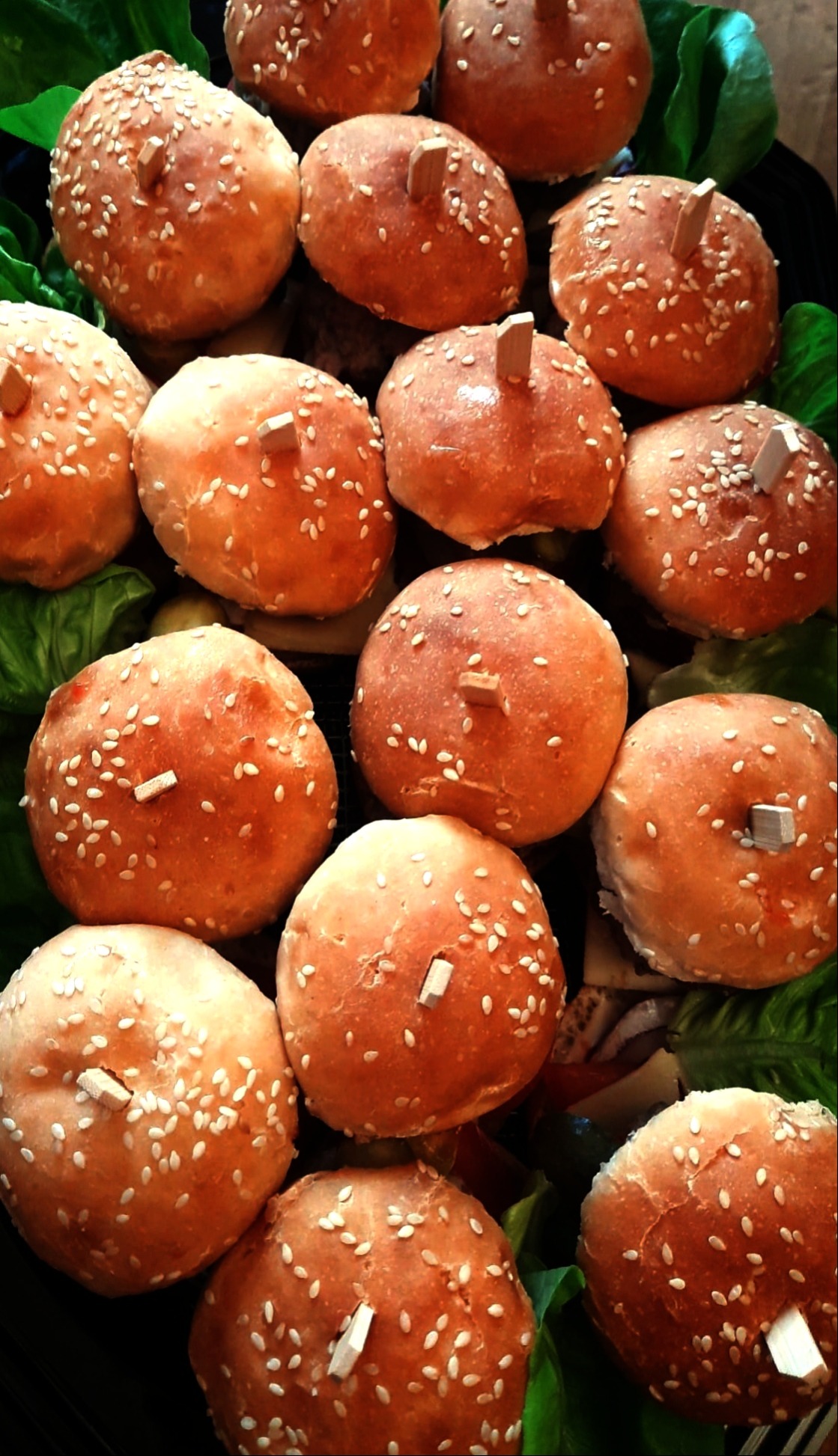 Mini bułeczki z sezamem i dekoracyjnymi wykałaczkami, ułożone na zielonych liściach sałaty. Idealne jako przekąska cateringowa na imprezę.