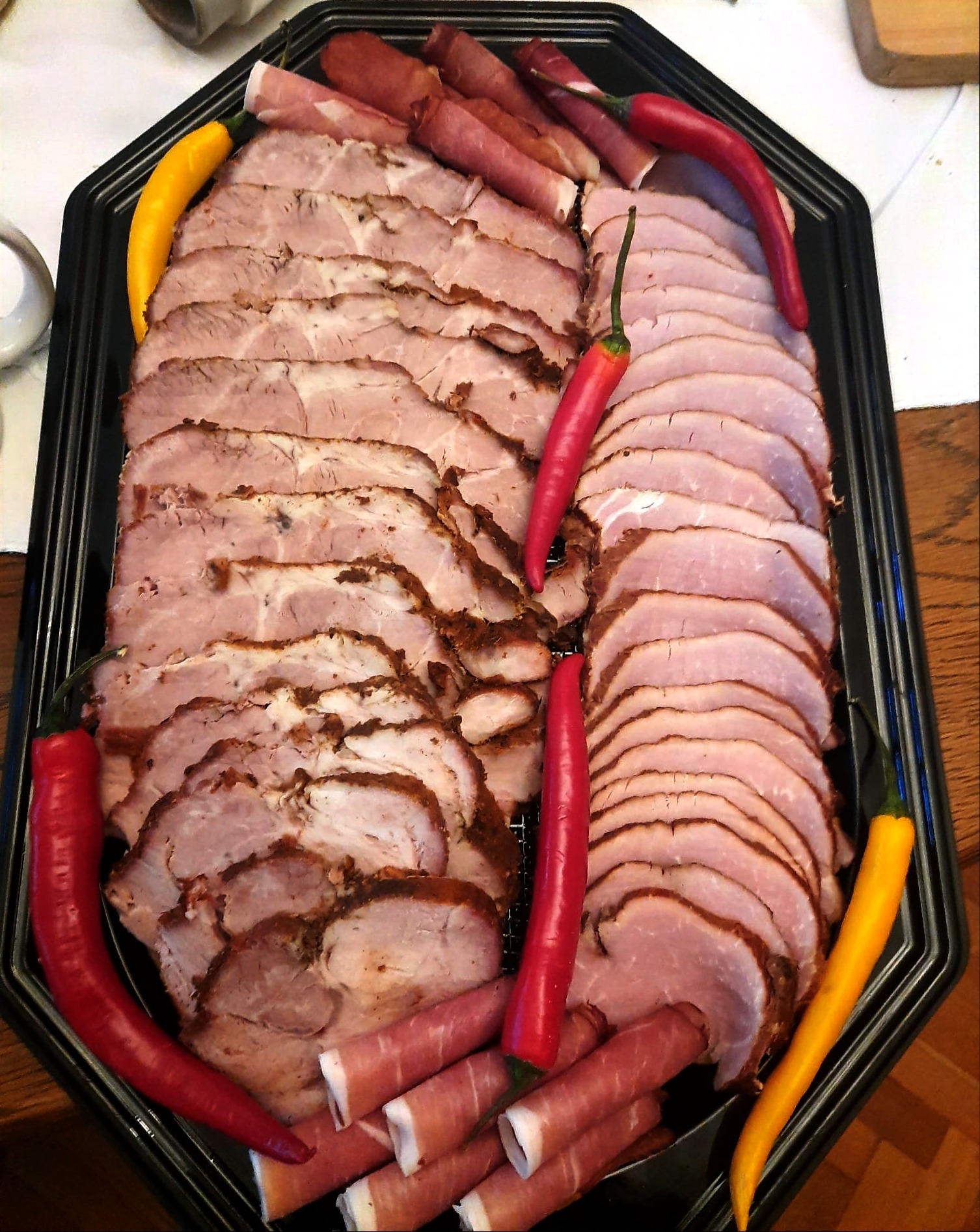 Elegancka taca z wędlinami: plastry pieczeni i szynki, dekorowane roladkami z szynki parmeńskiej i papryczkami chili w kolorach czerwonym i żółtym. Idealne na catering.
