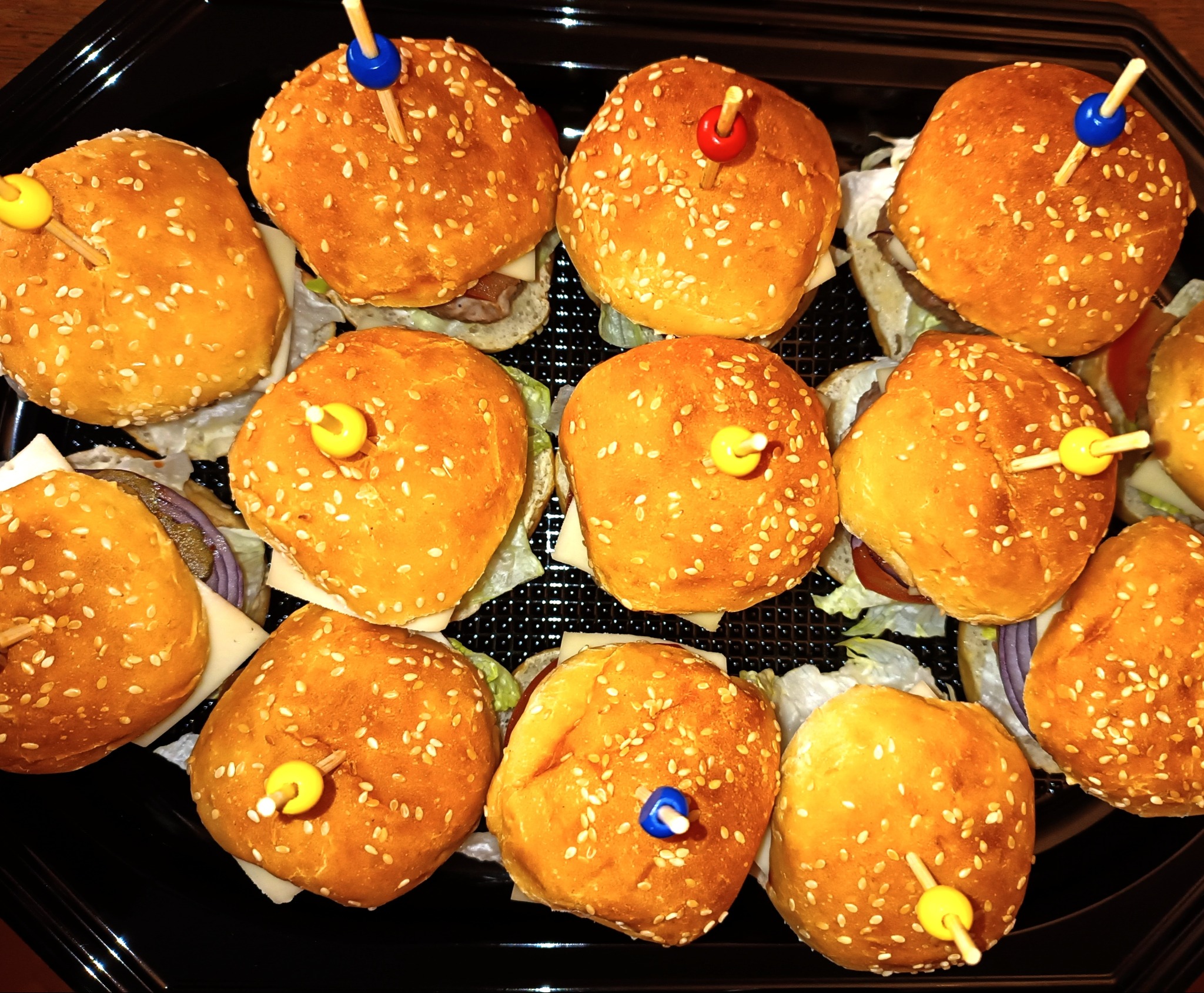 Apettyczne mini burgery z kolorowymi wykałaczkami na czarnej tacy, idealne na catering firmowy lub imprezę. Bułeczki posypane sezamem, widoczne warzywa i ser.