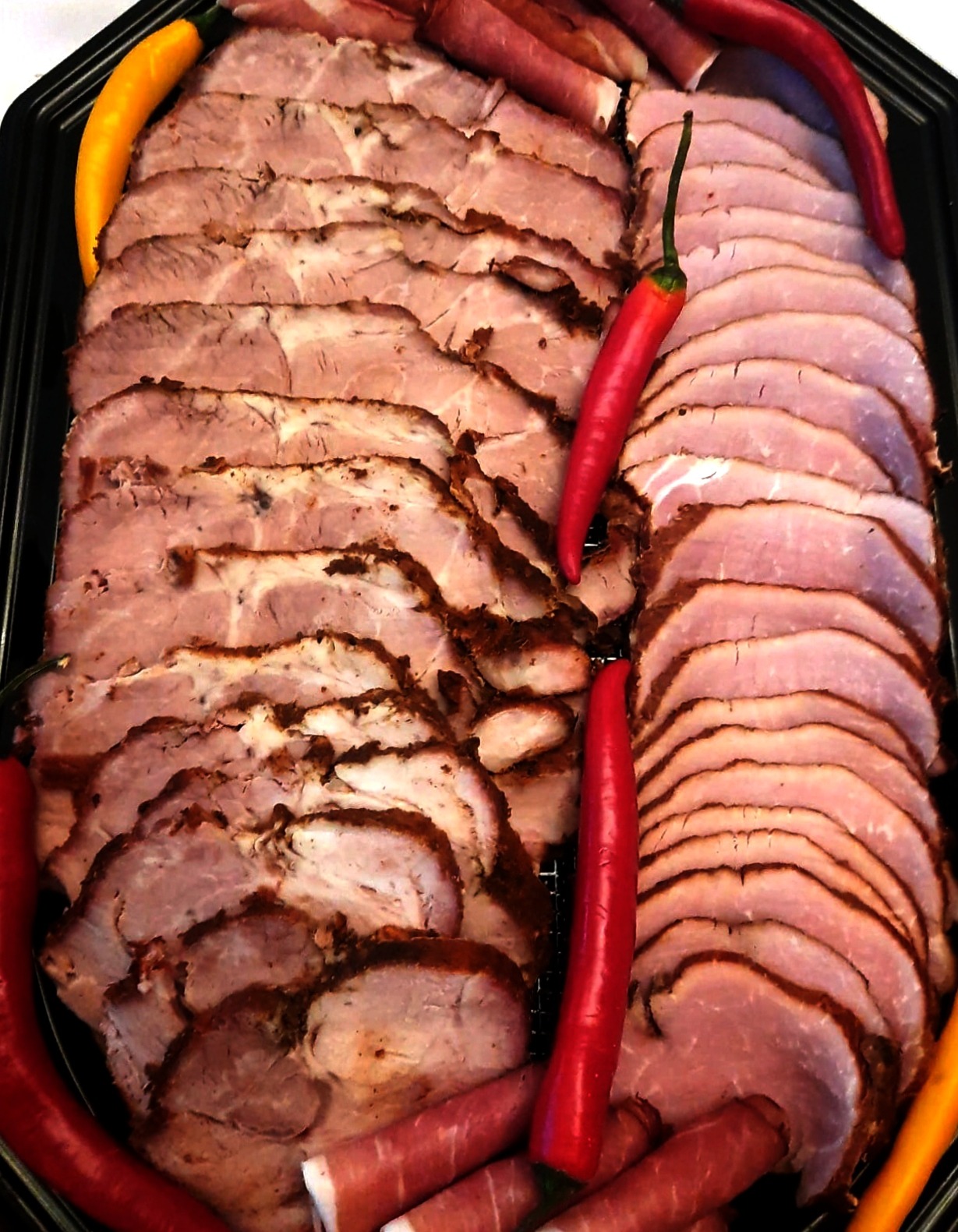 Czarny półmisek z układem wędlin: plastry pieczonego mięsa i szynki, dekorowane papryczkami chili i roladkami szynki parmeńskiej. Elegancki catering na imprezę.