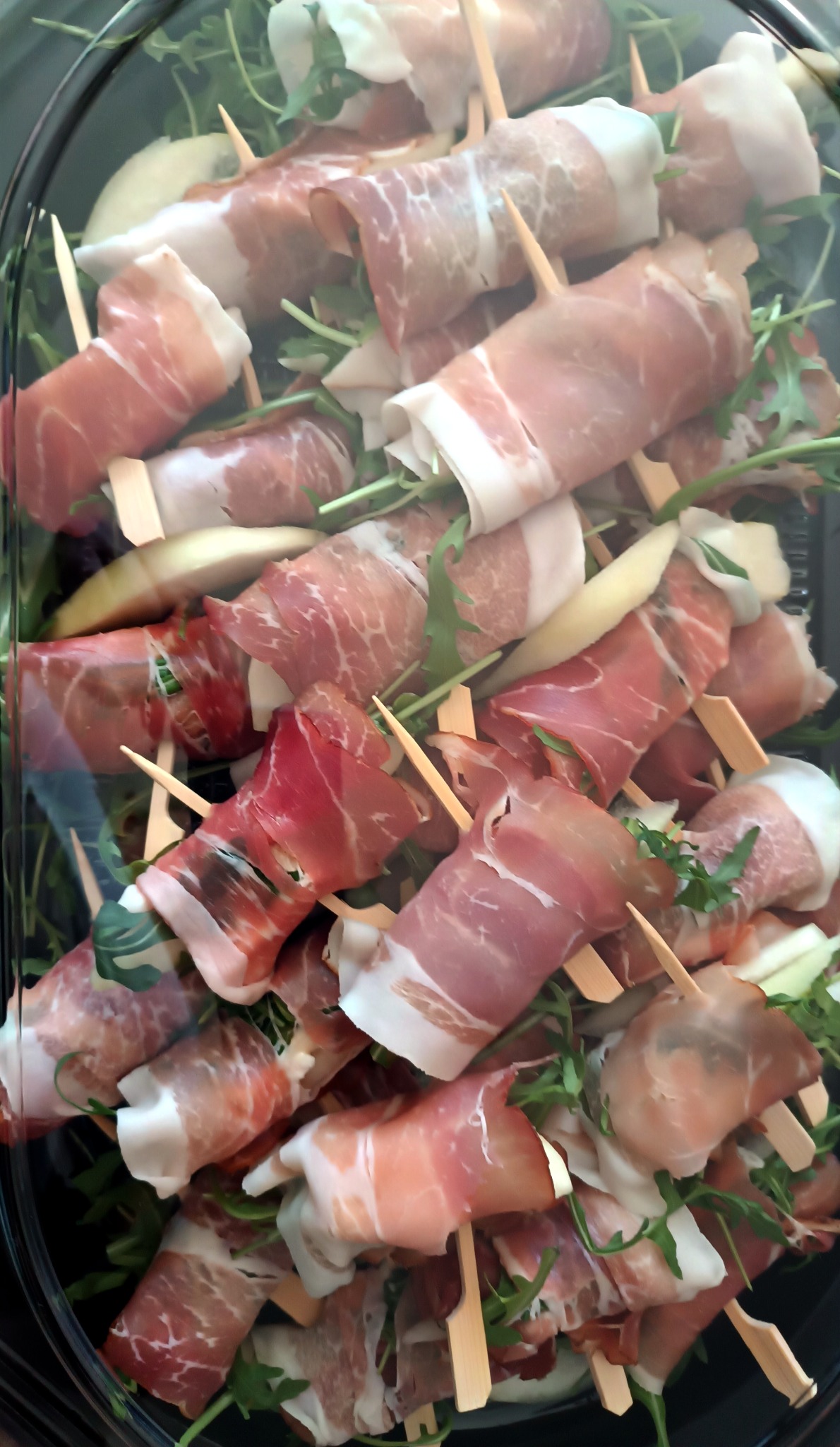 Eleganckie koreczki z szynki parmeńskiej i melona na rukoli, podane w czarnym, plastikowym pojemniku cateringowym. Idealne na imprezę firmową lub przyjęcie świąteczne.