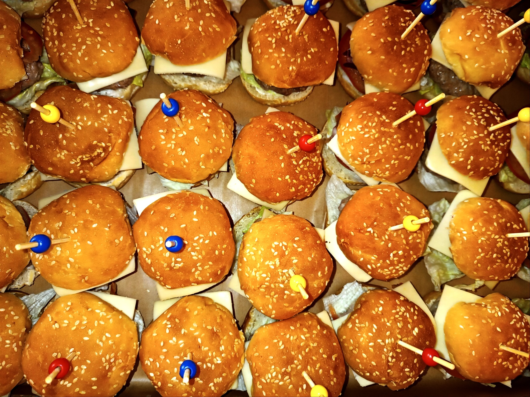 Mini burgery z serem i kolorowymi wykałaczkami, ułożone w rzędach. Widok z góry, ciepłe światło podkreśla złocisty kolor bułek posypanych sezamem.