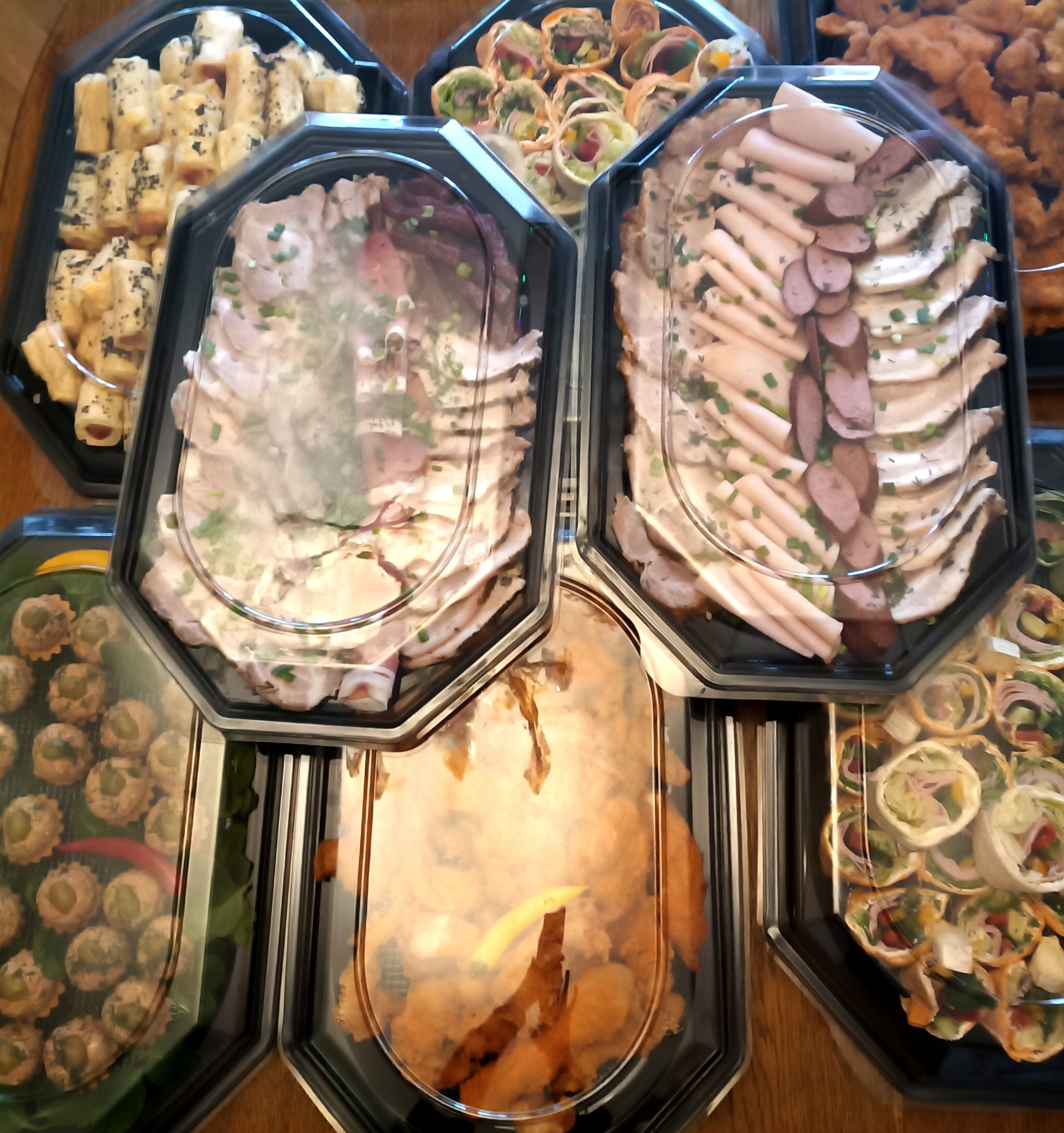 Zestaw różnorodnych przekąsek cateringowych w czarnych pojemnikach z przezroczystymi pokrywkami, ułożonych na drewnianym stole. Widoczne roladki, tartaletki i wędliny.