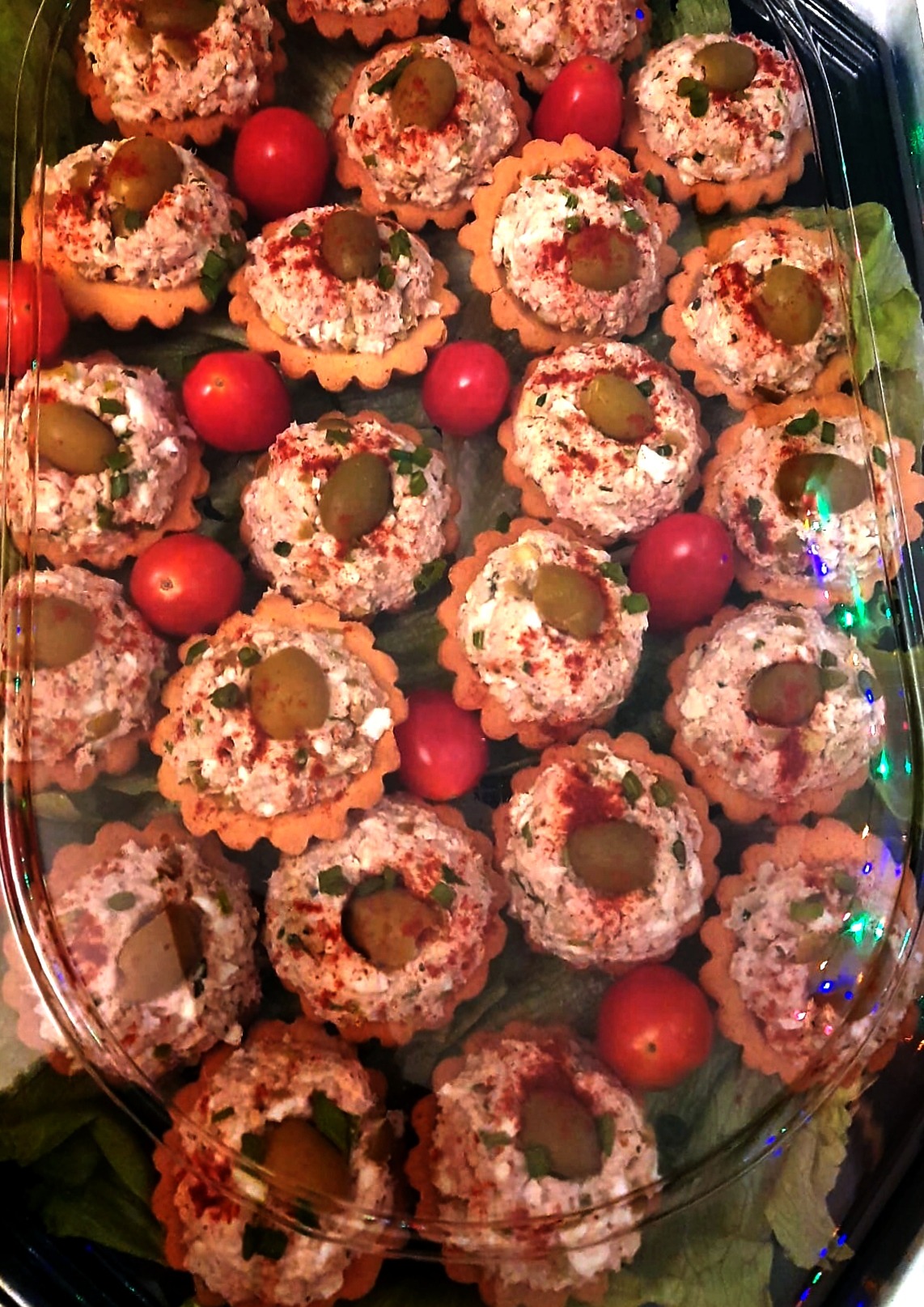 Tarty z pastą, oliwką i papryką, otoczone pomidorkami koktajlowymi na zielonych liściach, w przezroczystym opakowaniu cateringowym z delikatnym oświetleniem.