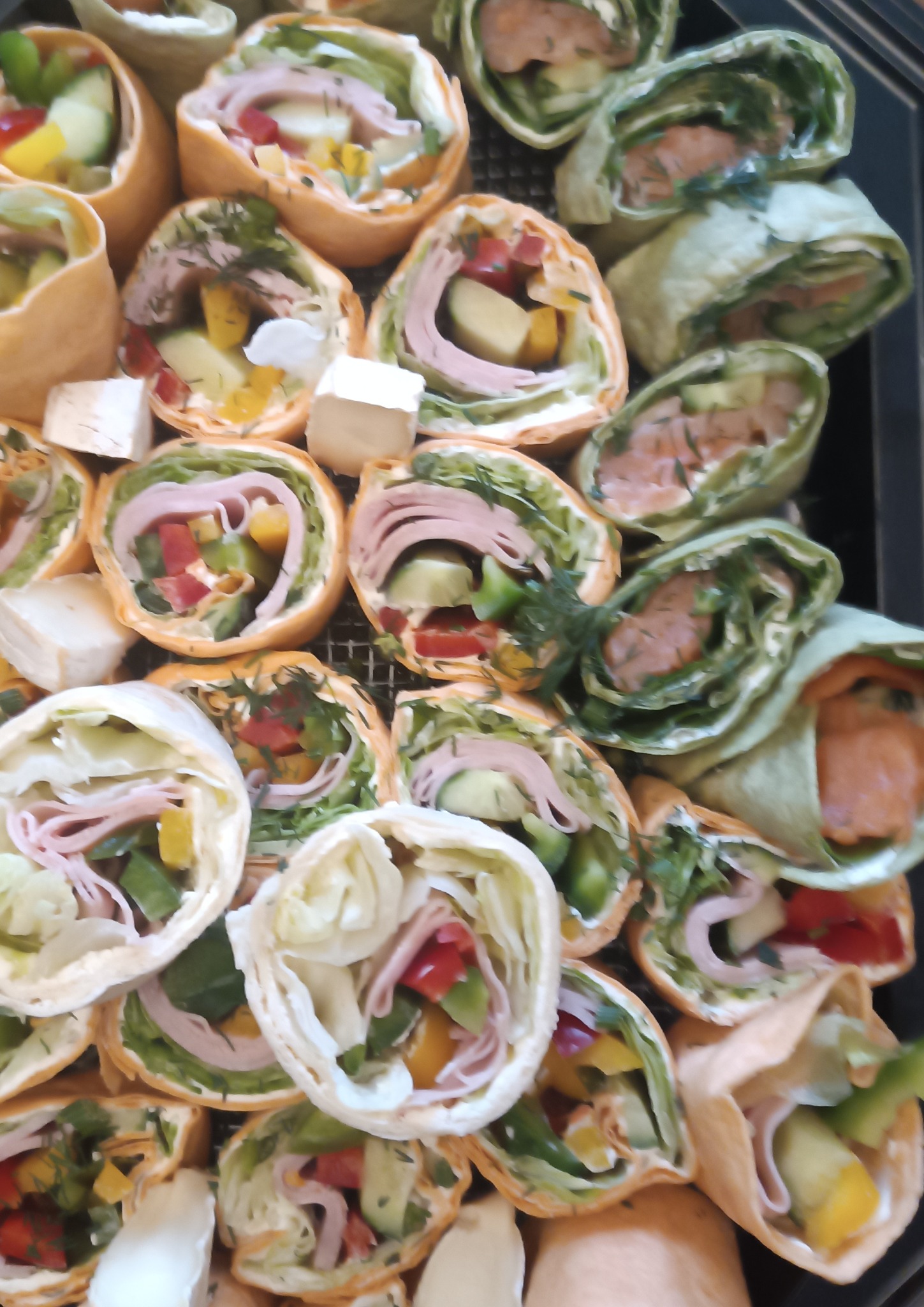 Kolorowe tortille z łososiem, szynką i warzywami, ułożone na czarnej tacy cateringowej. Widoczne kawałki sera brie. Różnorodność smaków i barw.