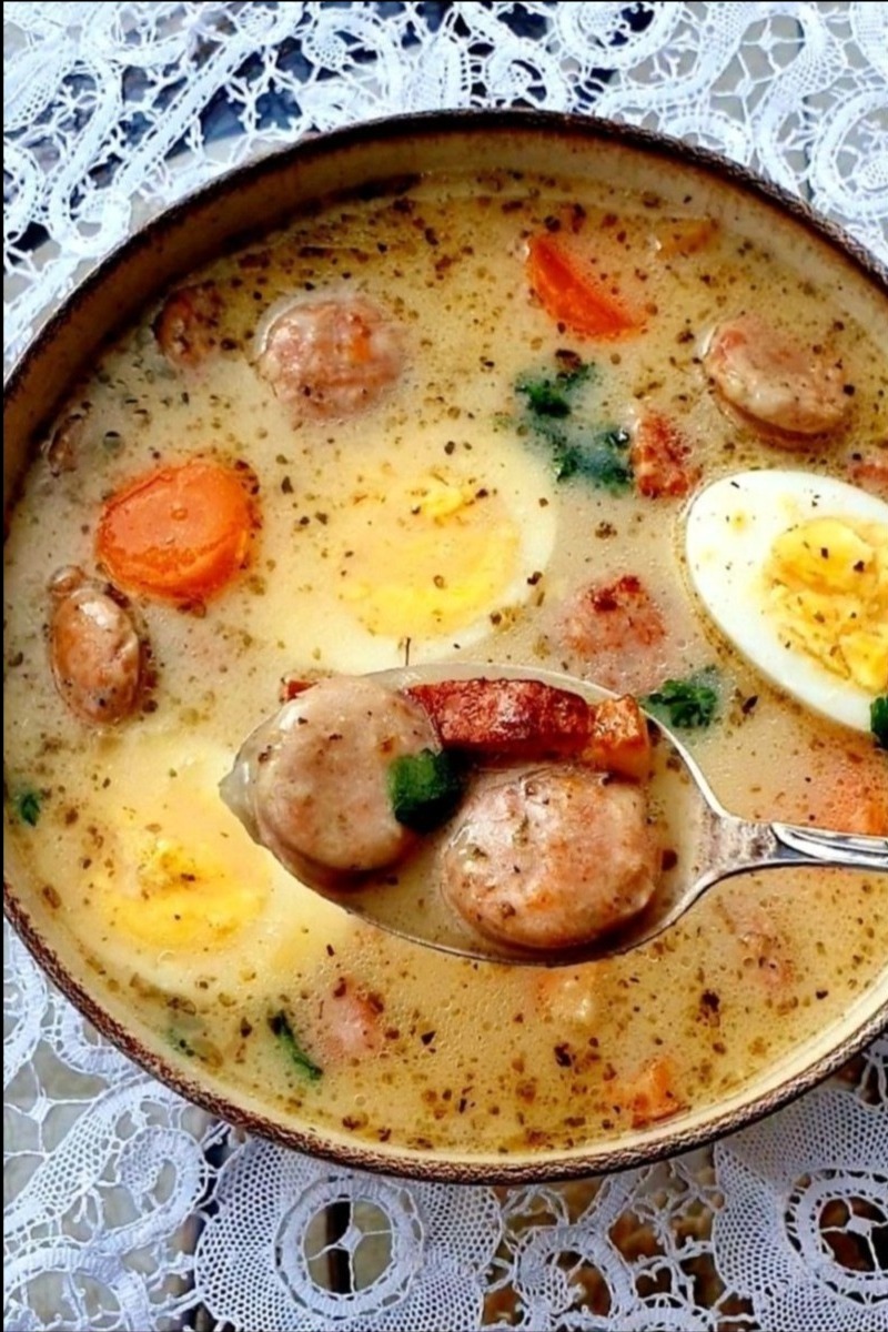 Żurek w ceramicznej misce, z jajkiem, kiełbasą i marchewką, podawany na łyżce. Dekoracyjny obrus w tle. Tradycyjna zupa w zbliżeniu, gotowa do spożycia.