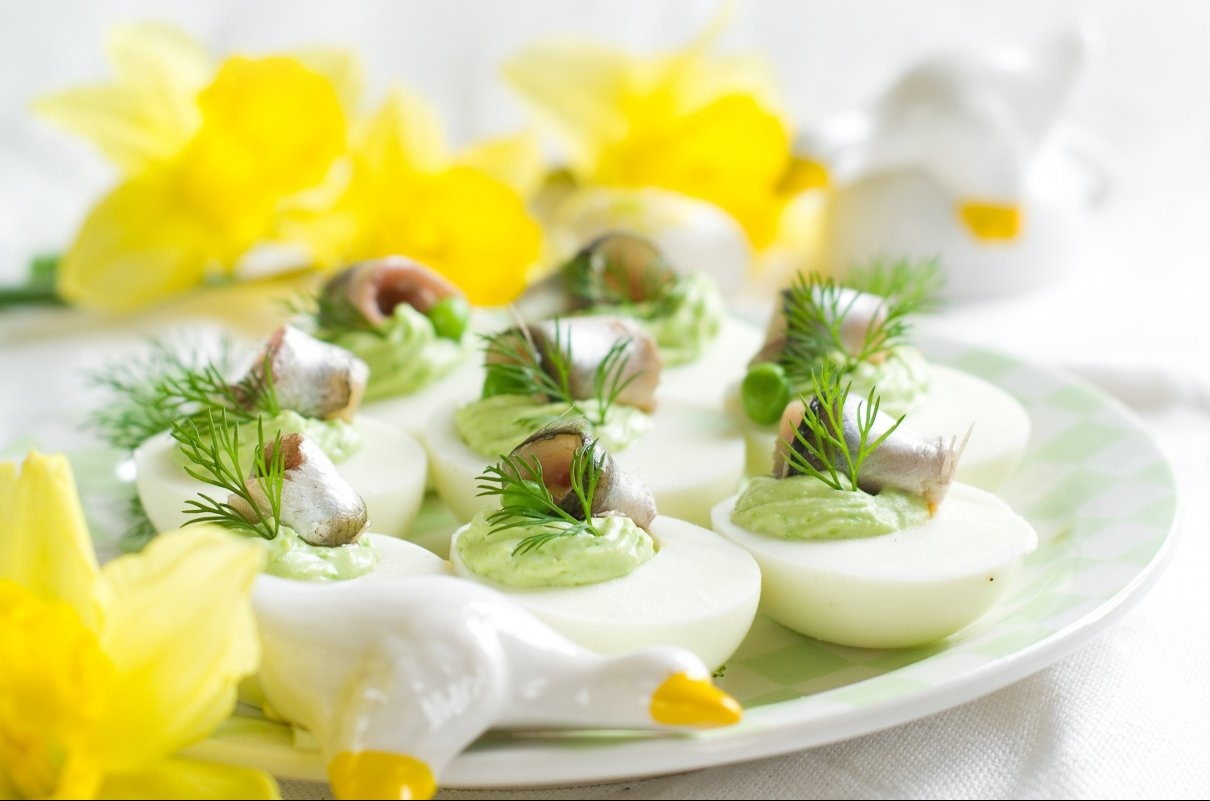 Dekoracyjne jajka faszerowane pastą, sardynką i koperkiem na talerzu w kratkę, z żółtymi kwiatami i figurką kaczki w tle. Delikatne, jasne oświetlenie.