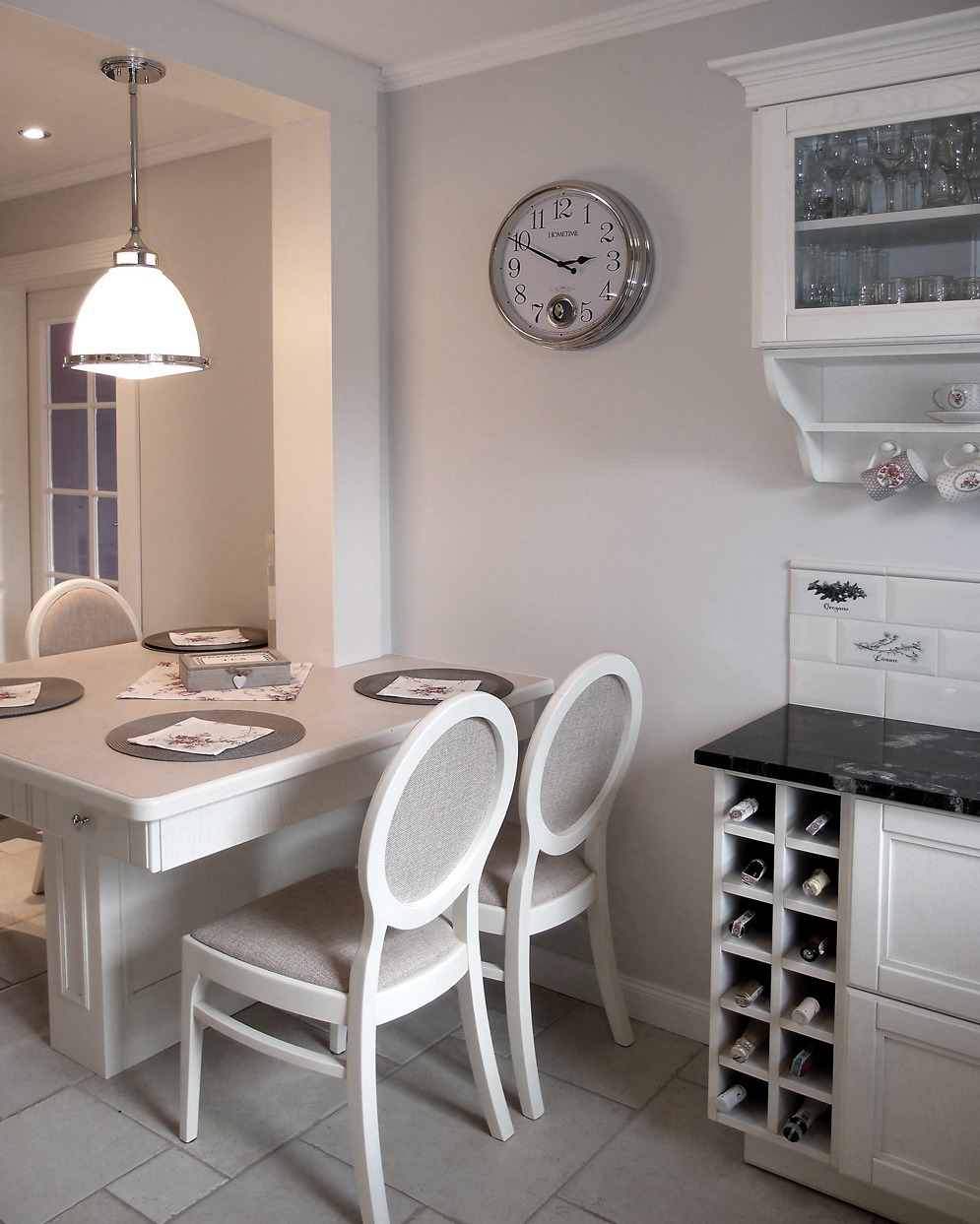 Jasna jadalnia z białymi meblami: stół z krzesłami, szafka z półkami na wino, zegar ścienny i lampa wisząca, utrzymana w stylu klasycznym.