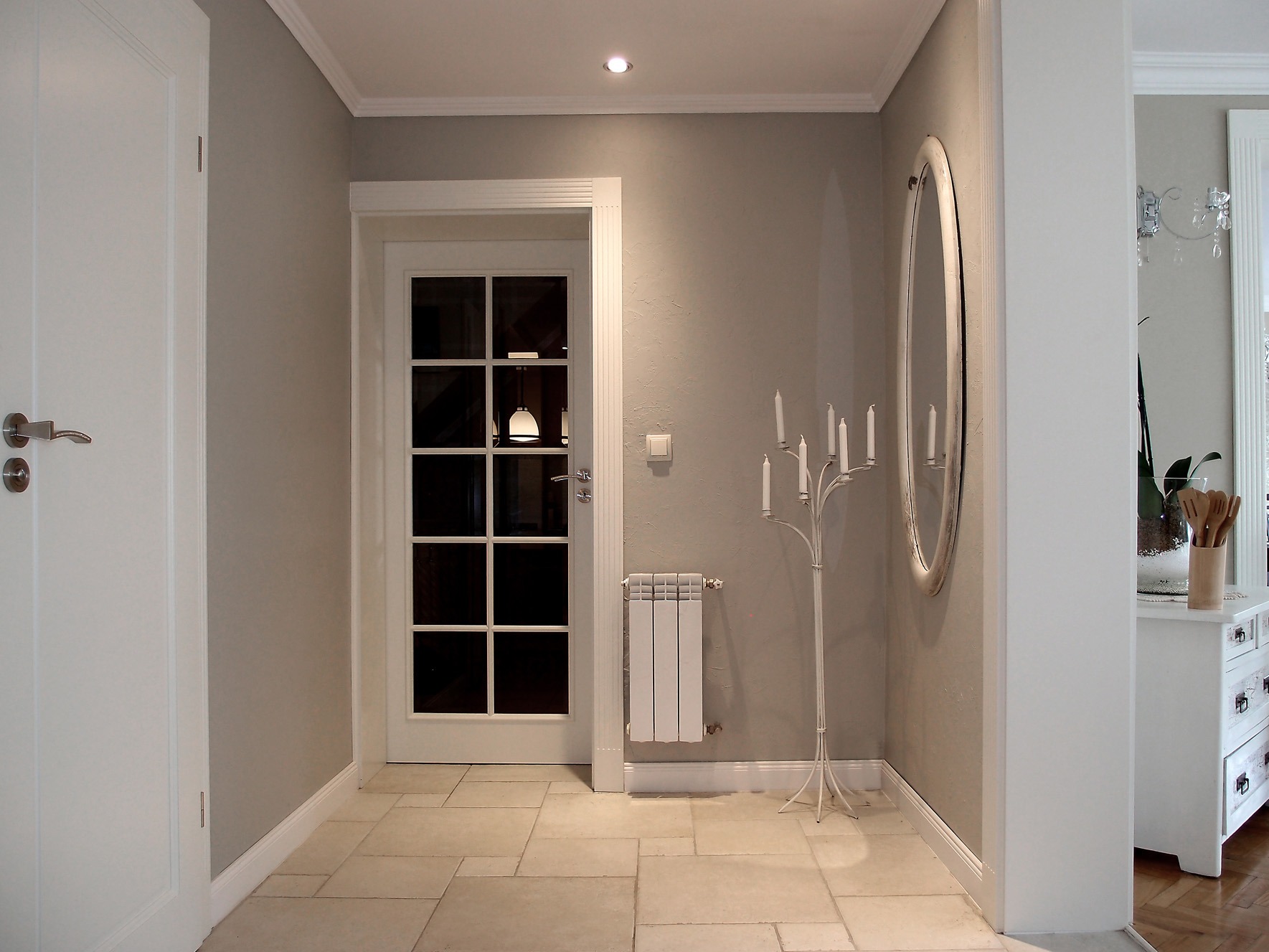 Neutralna aranżacja przedpokoju z białymi drzwiami, grzejnikiem, lustrem w owalnej ramie i świecznikiem podłogowym na siedem świec.