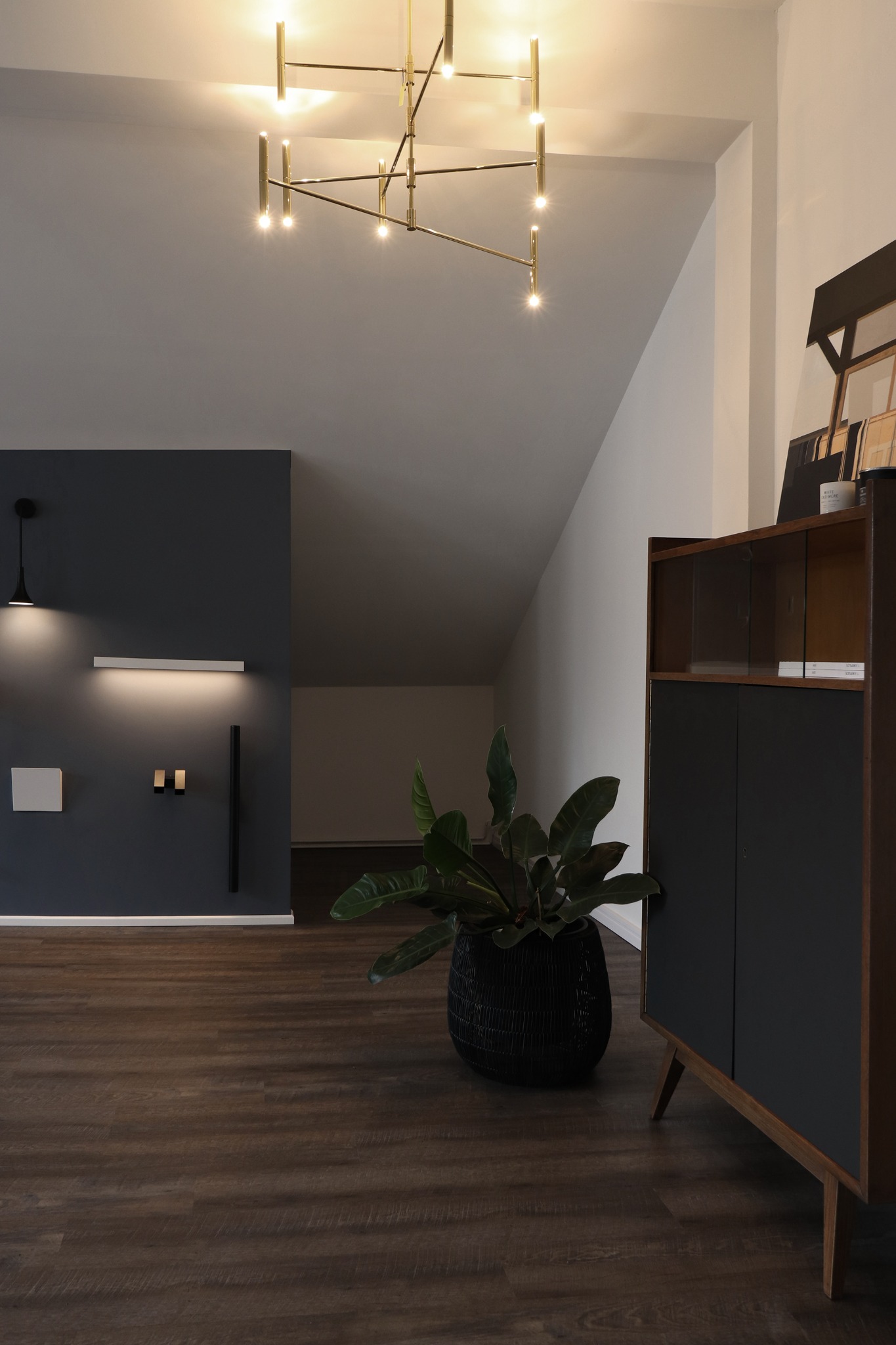 Minimalistyczne wnętrze z ciemną ścianą, oświetleniem punktowym, drewnianą podłogą i szafką vintage z rośliną doniczkową, oświetlone nowoczesną, złotą lampą sufitową.