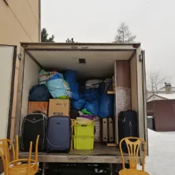 Wnętrze furgonetki wypełnionej po brzegi spakowanymi rzeczami, w tym walizkami, kartonami i workami, z dwoma żółtymi krzesłami opartymi o tylną klapę na tle zimowego krajobrazu.