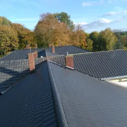 Widok z góry na ciemnoszary dach z dachówki ceramicznej z widocznymi kominami z czerwonej cegły, na tle jesiennego krajobrazu z drzewami o różnorodnych barwach.