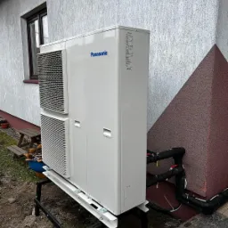 Biała pompa ciepła Panasonic na czarnym metalowym stelażu, zainstalowana przy ścianie budynku z widocznymi rurami w izolacji termicznej.